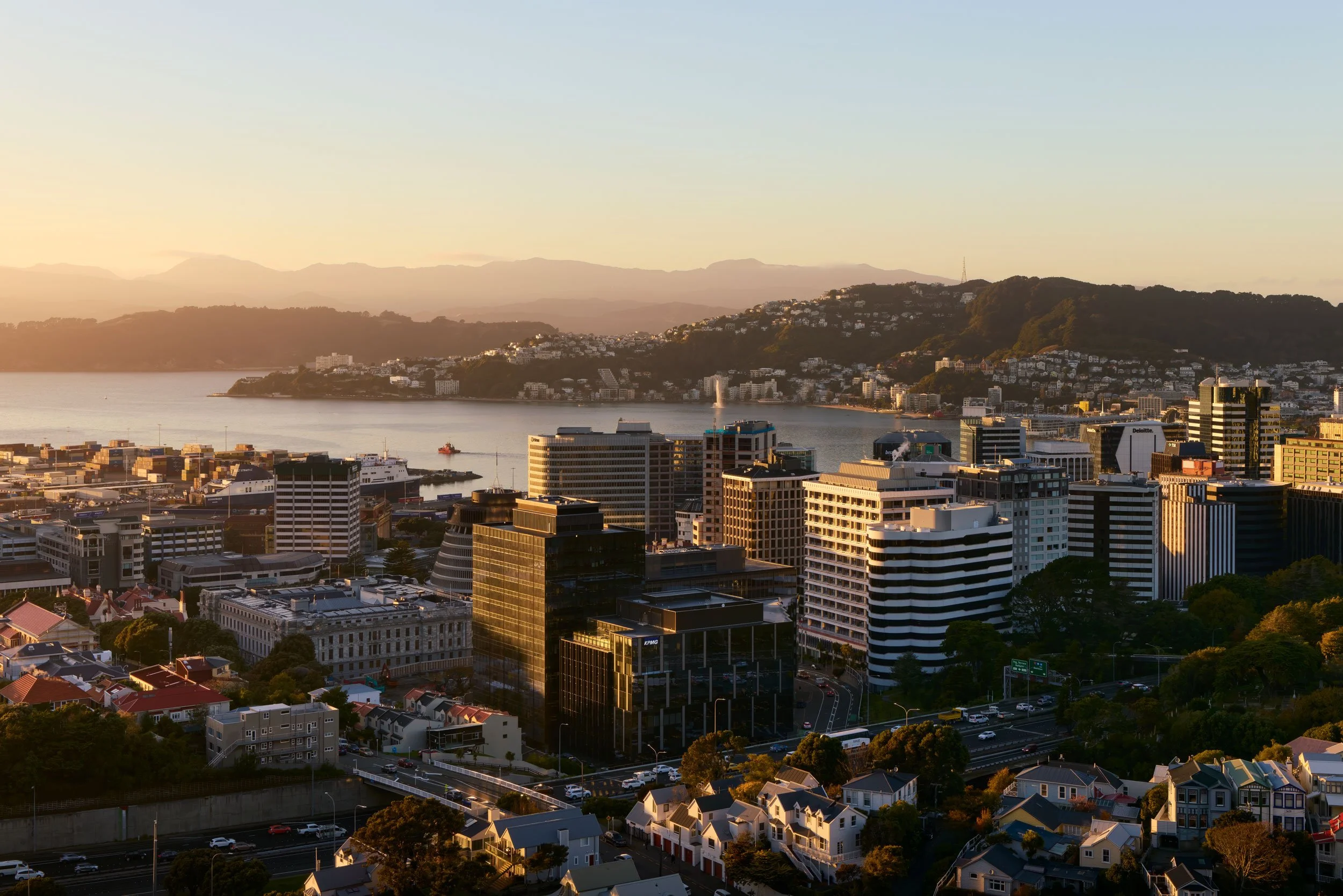 A cityscape at sunset featuring tall modern buildings near a body of water, with a hillside dotted with houses in the background.