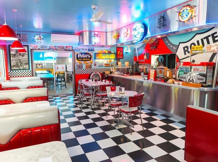 Interior of a retro American diner with checkered black and white floor, red and white booths, small tables, and a counter with seating. Neon signs and Route 66 decor adorn the walls.