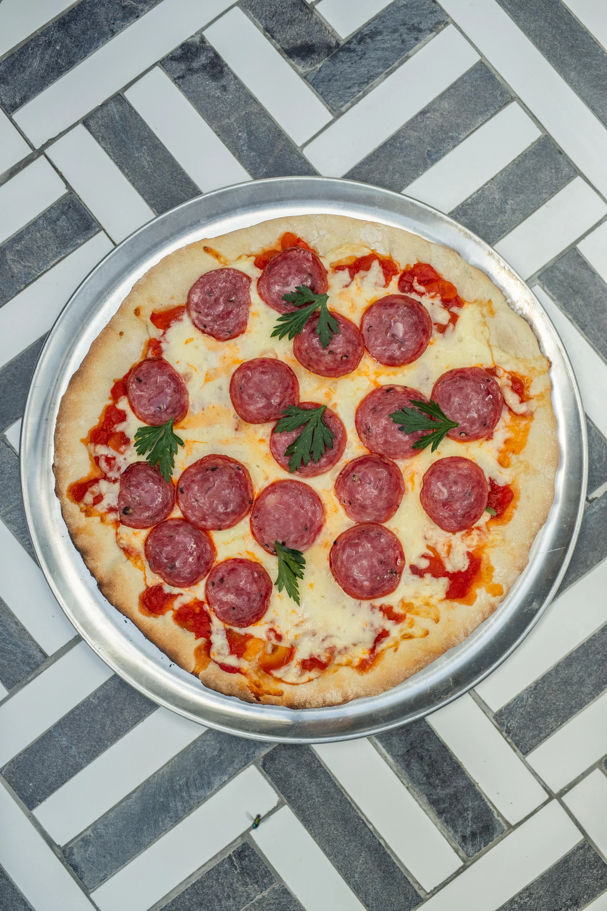 A pepperoni pizza with some parsley garnishes on a silver tray, placed on a checked black and white tiled floor.