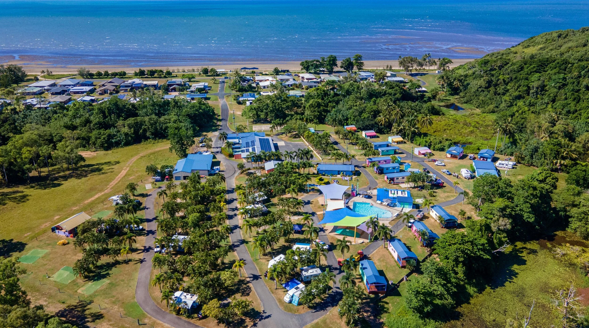 Aerial view of a coastal campground with colorful cabins, a swimming pool, and lush greenery, overlooking a beach and the ocean.