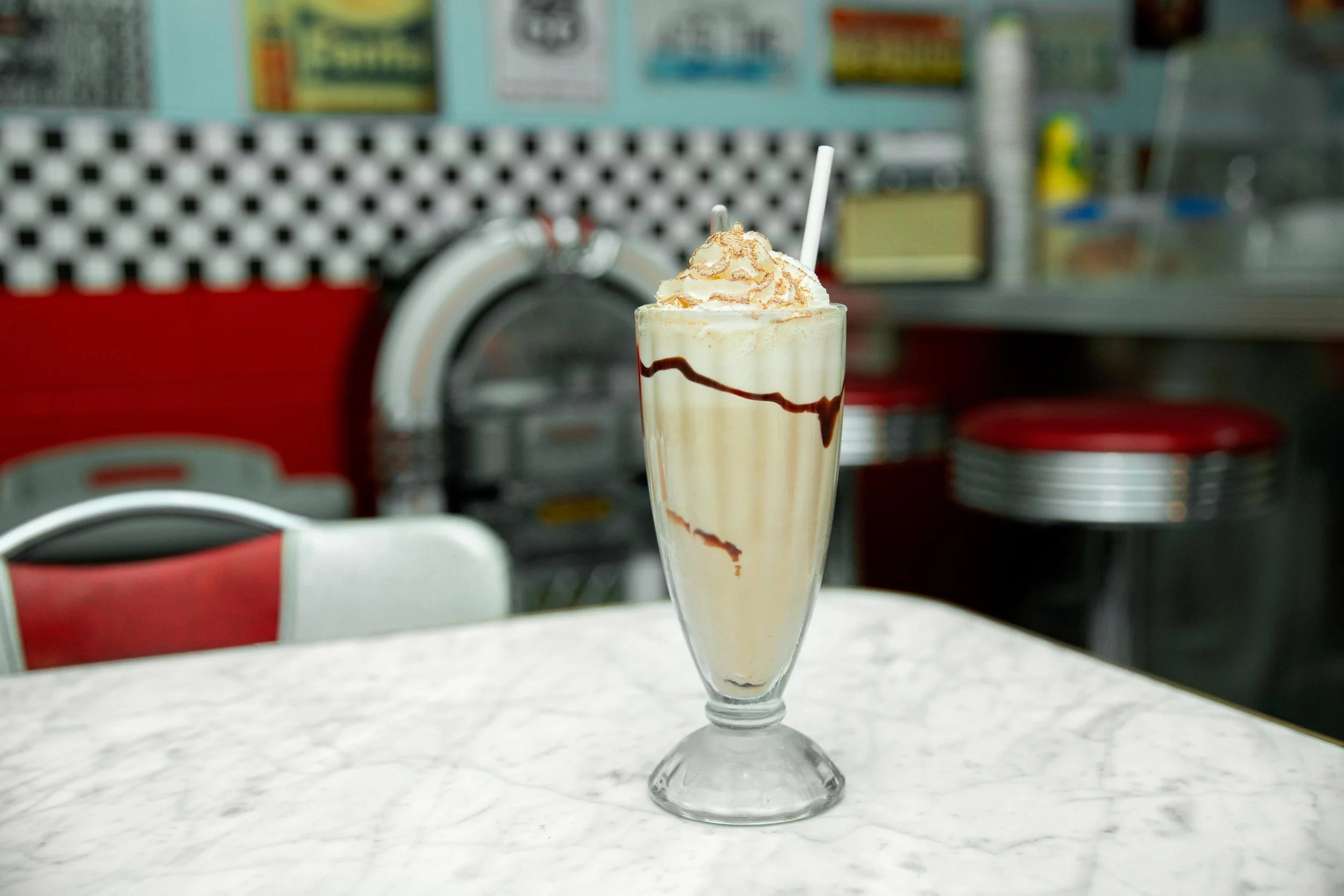 A tall glass of milkshake topped with whipped cream and caramel drizzle, with a straw, on a white countertop in a retro diner setting.