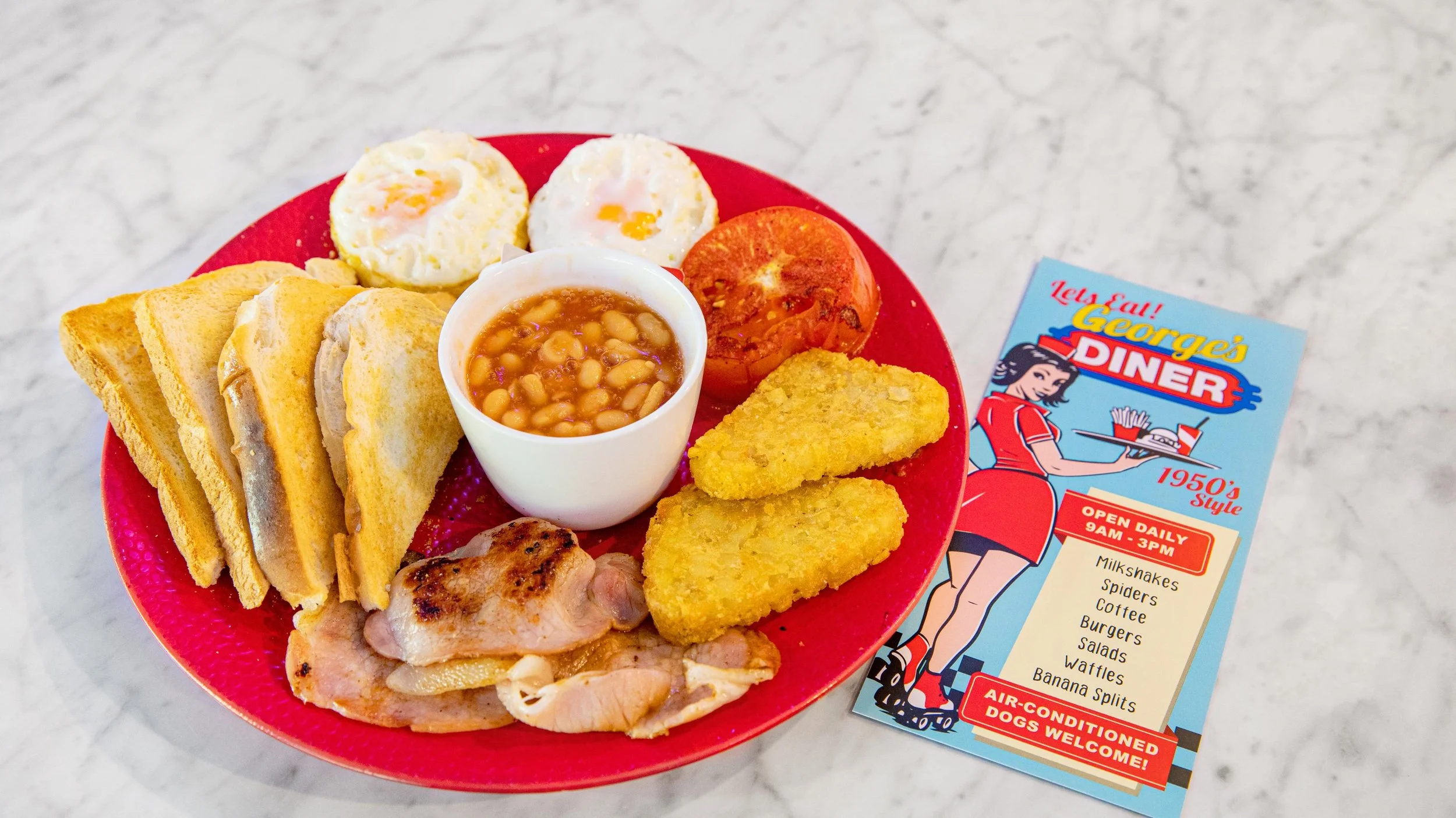 A red plate with eggs, toast, baked beans, hash browns, grilled chicken, and cooked ham. There is a flyer for George's Diner and a marble surface in the background.