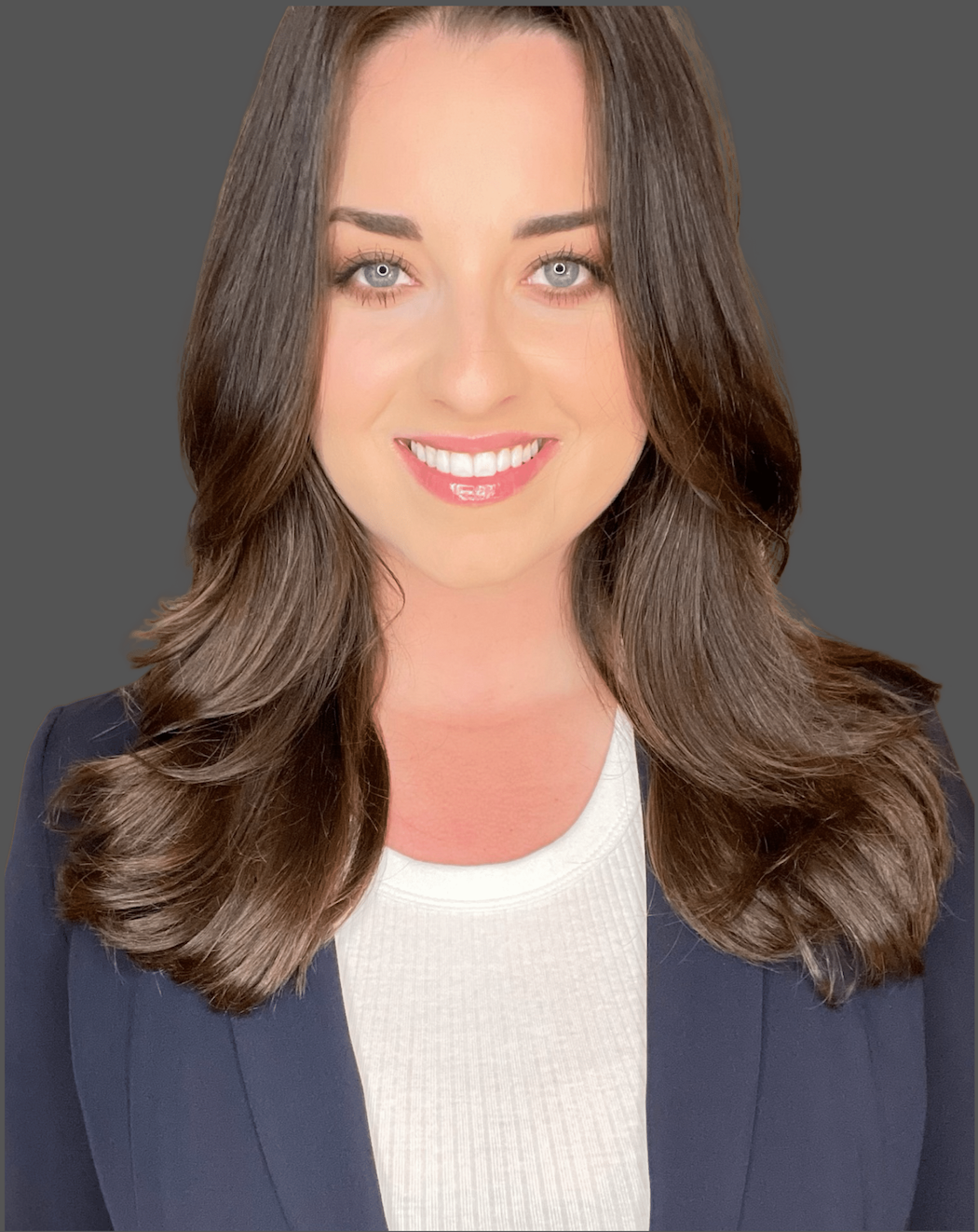 Headshot of a woman with long, wavy brown hair, blue eyes, and light skin, smiling, wearing a white shirt and dark blazer against a gray background.