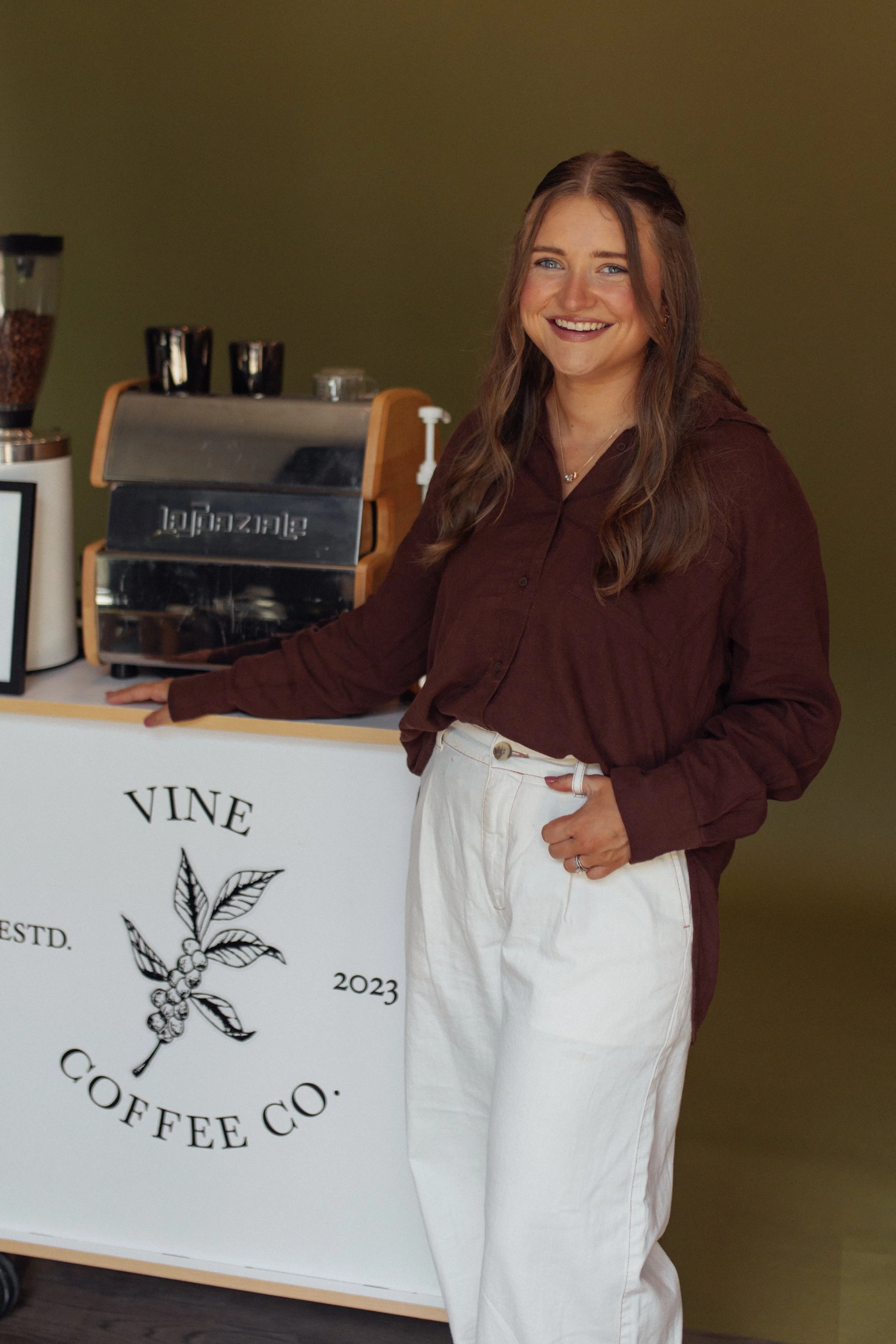 A woman with long brown hair, wearing a brown shirt and white pants, smiling and standing beside a coffee bar with a logo of a coffee plant and the words "Vine Coffee Co.".