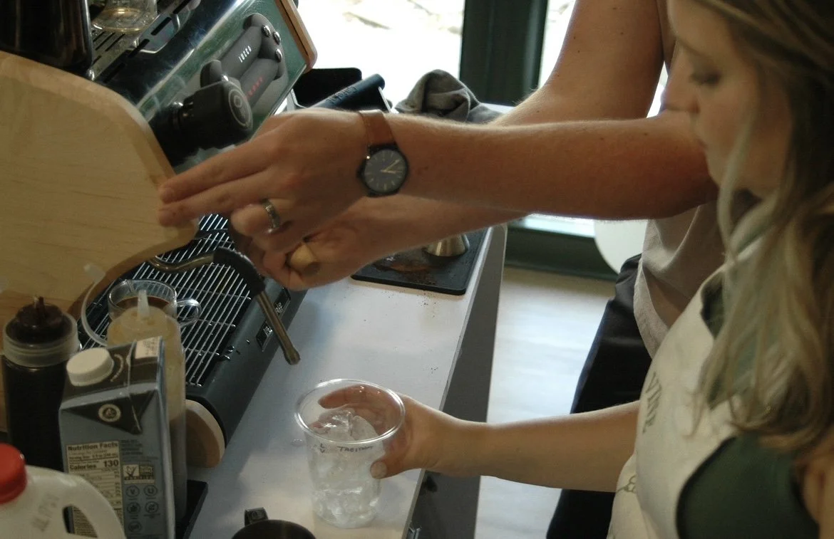 Person pouring coffee into a cup while a young girl holds a glass of ice water nearby in a kitchen.