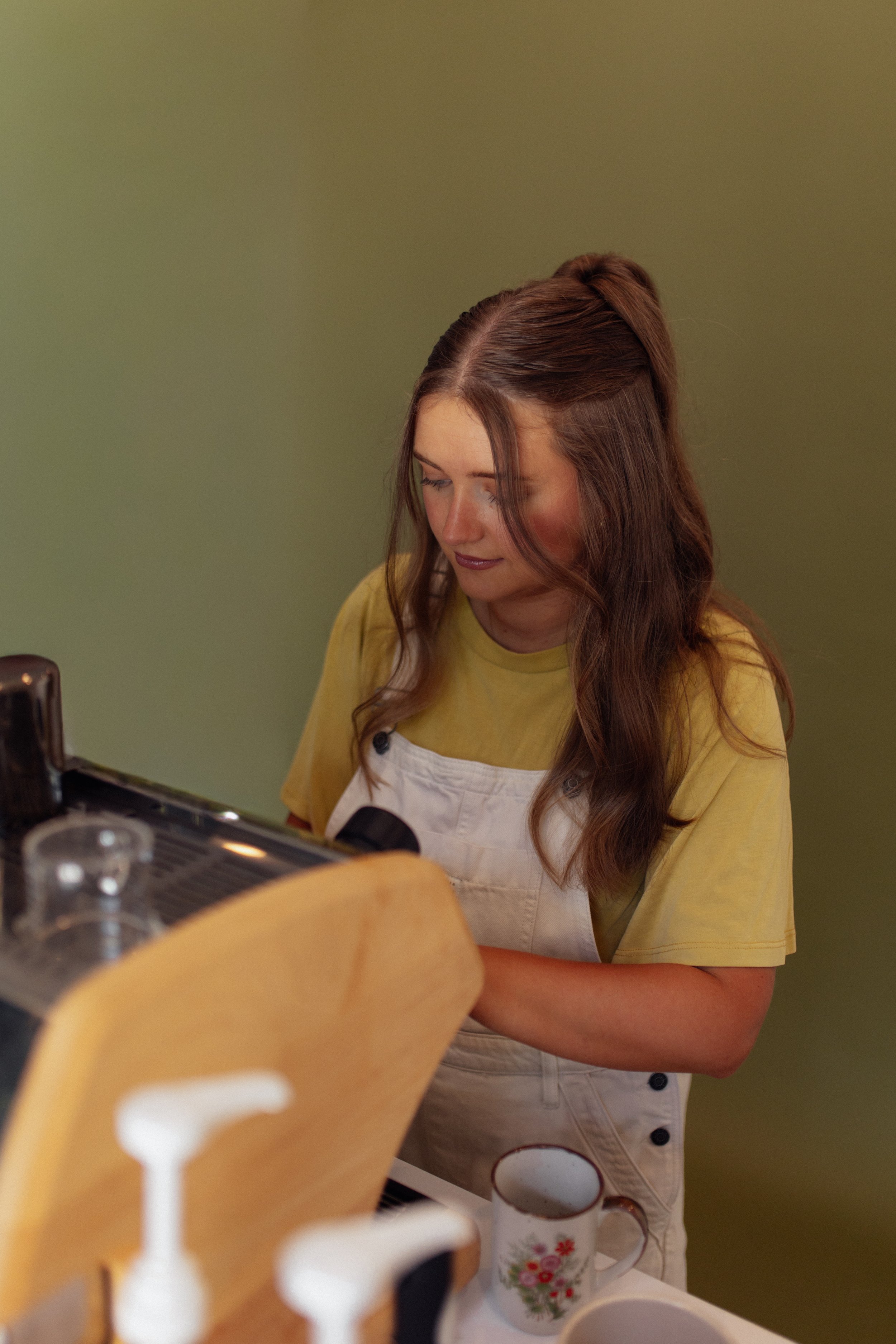 A woman with long brown hair, wearing a yellow shirt and white apron, standing in a kitchen against a green wall, preparing coffee or tea.