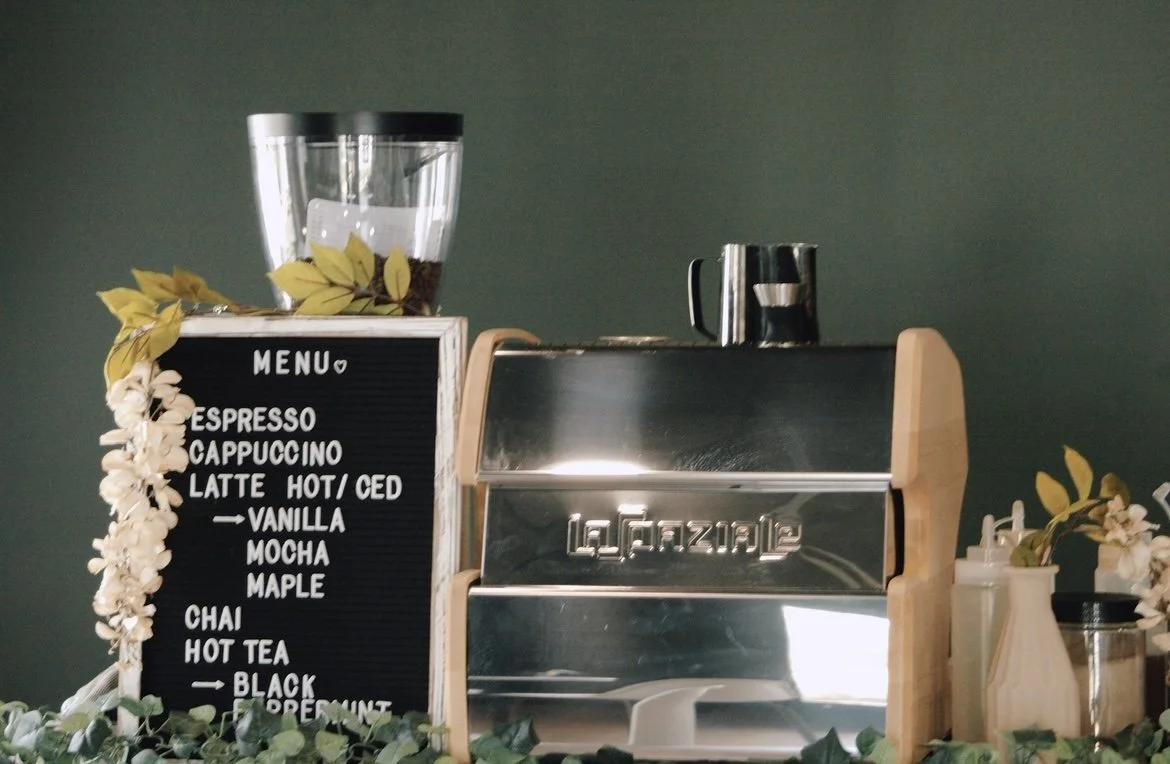 Coffee shop espresso machine with a black menu board listing coffee options, decorated with white flowers and green foliage.