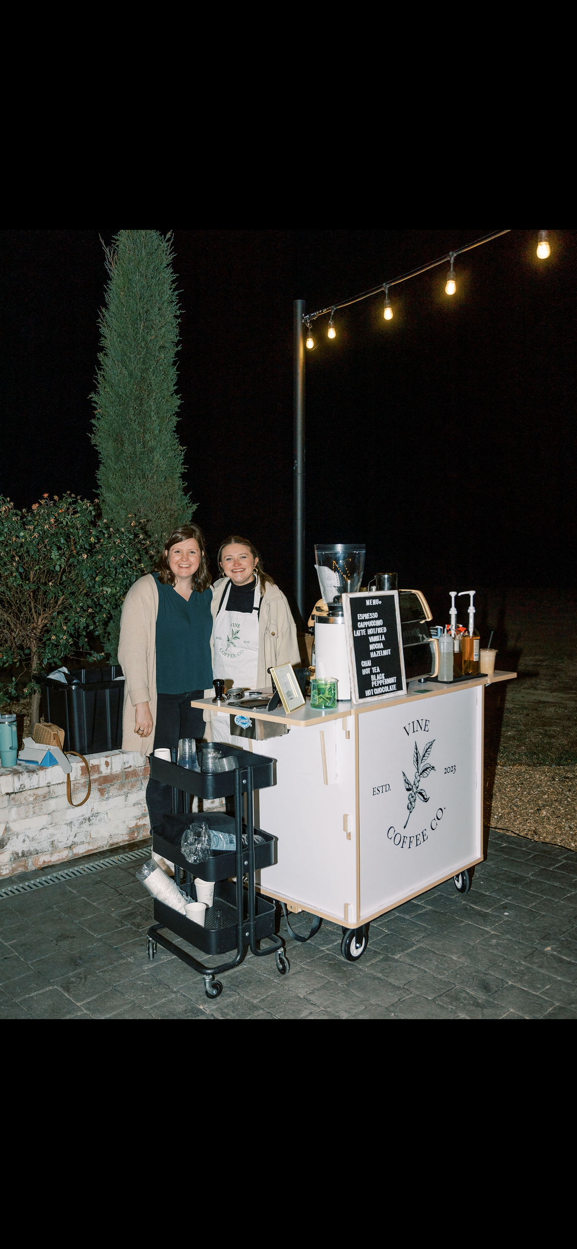 Two women standing behind a mobile coffee stand at night, smiling for the camera. The stand displays a menu with coffee options and is decorated with a logo of a leaf and the words "Vine Coffee Co." A black cart with cups and supplies is adjacent to 