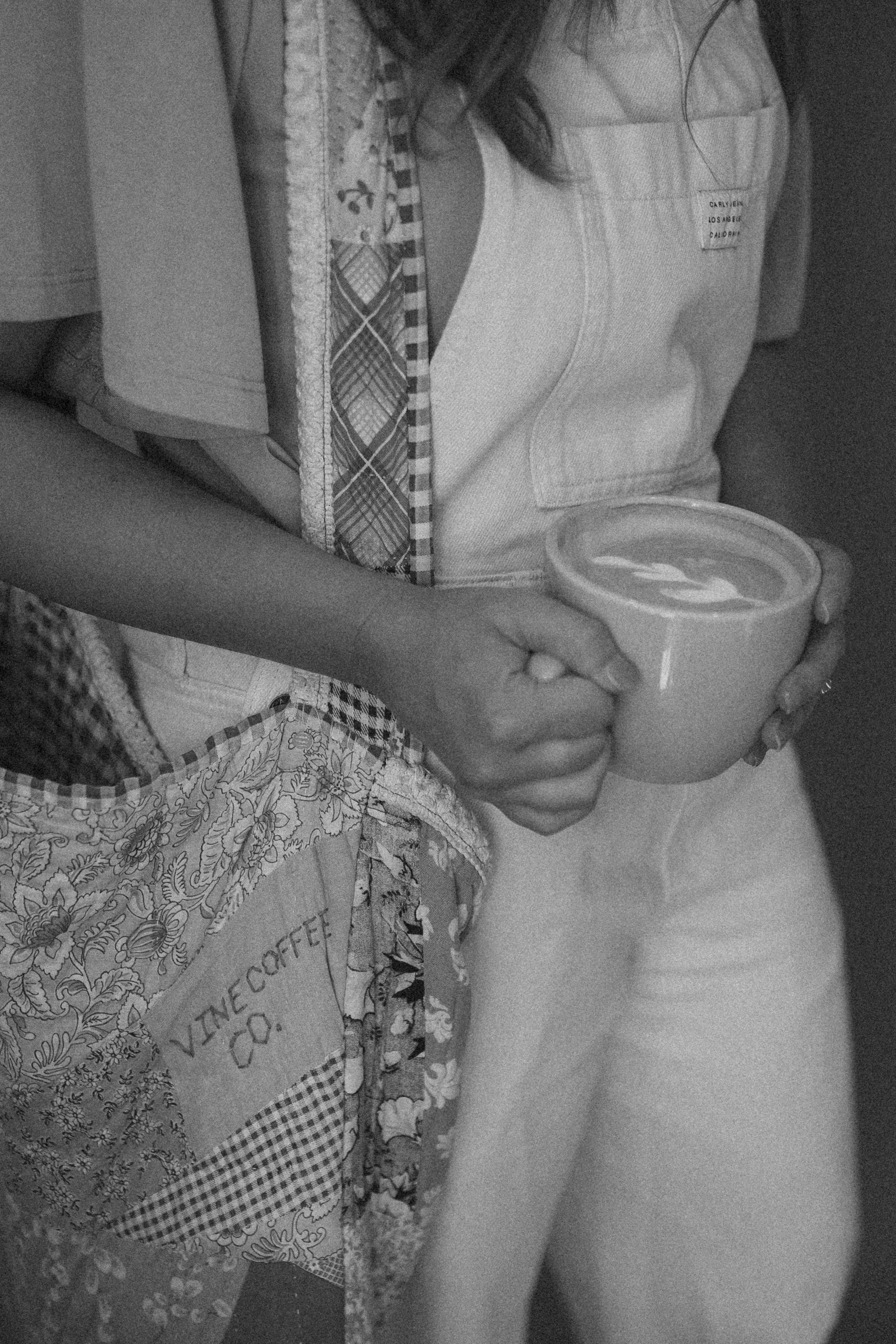 A person sitting down, wearing a light-colored shirt and patterned patchwork pants. The person's hand is holding a cup with a design on it, and part of their arm and hand are visible.