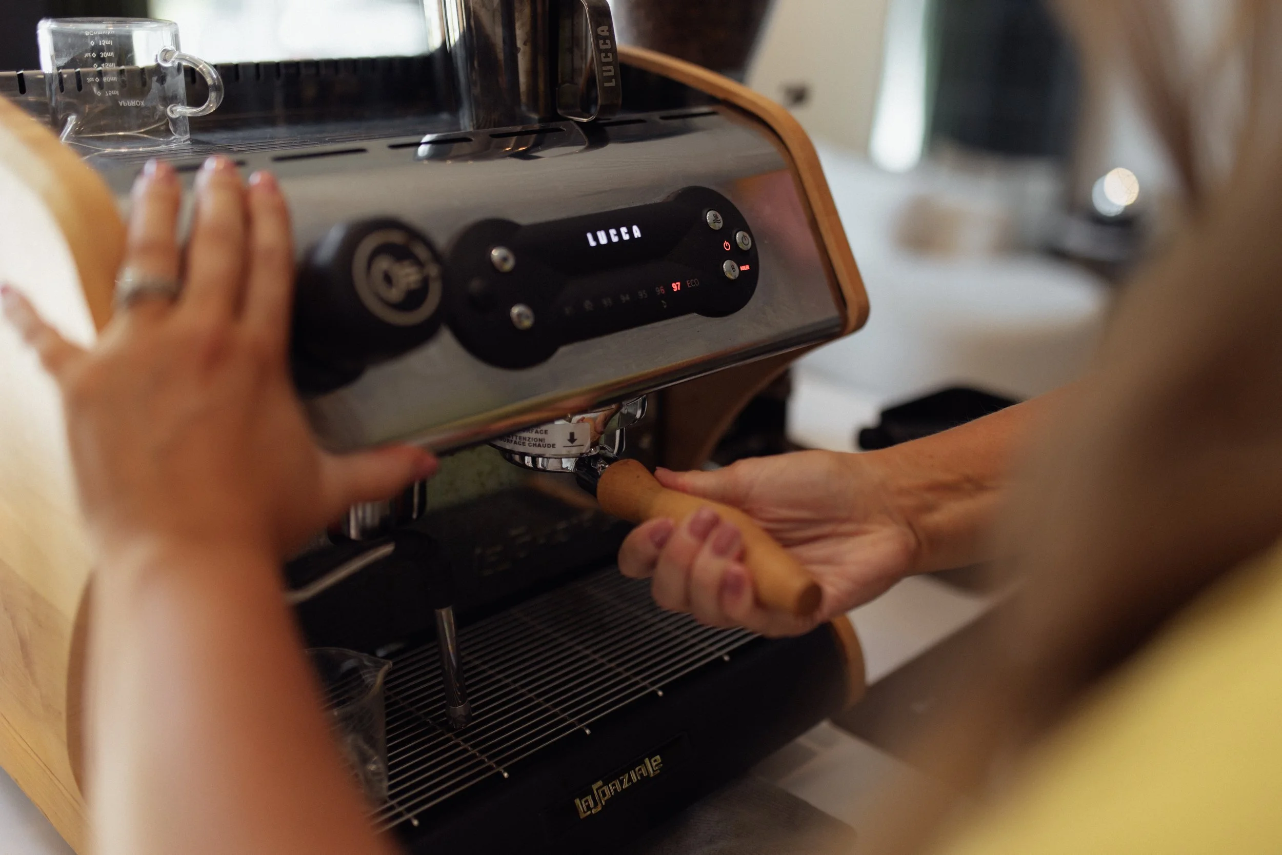 A person pressing a button on a Lucea espresso machine, with a hand holding a wooden stick below.