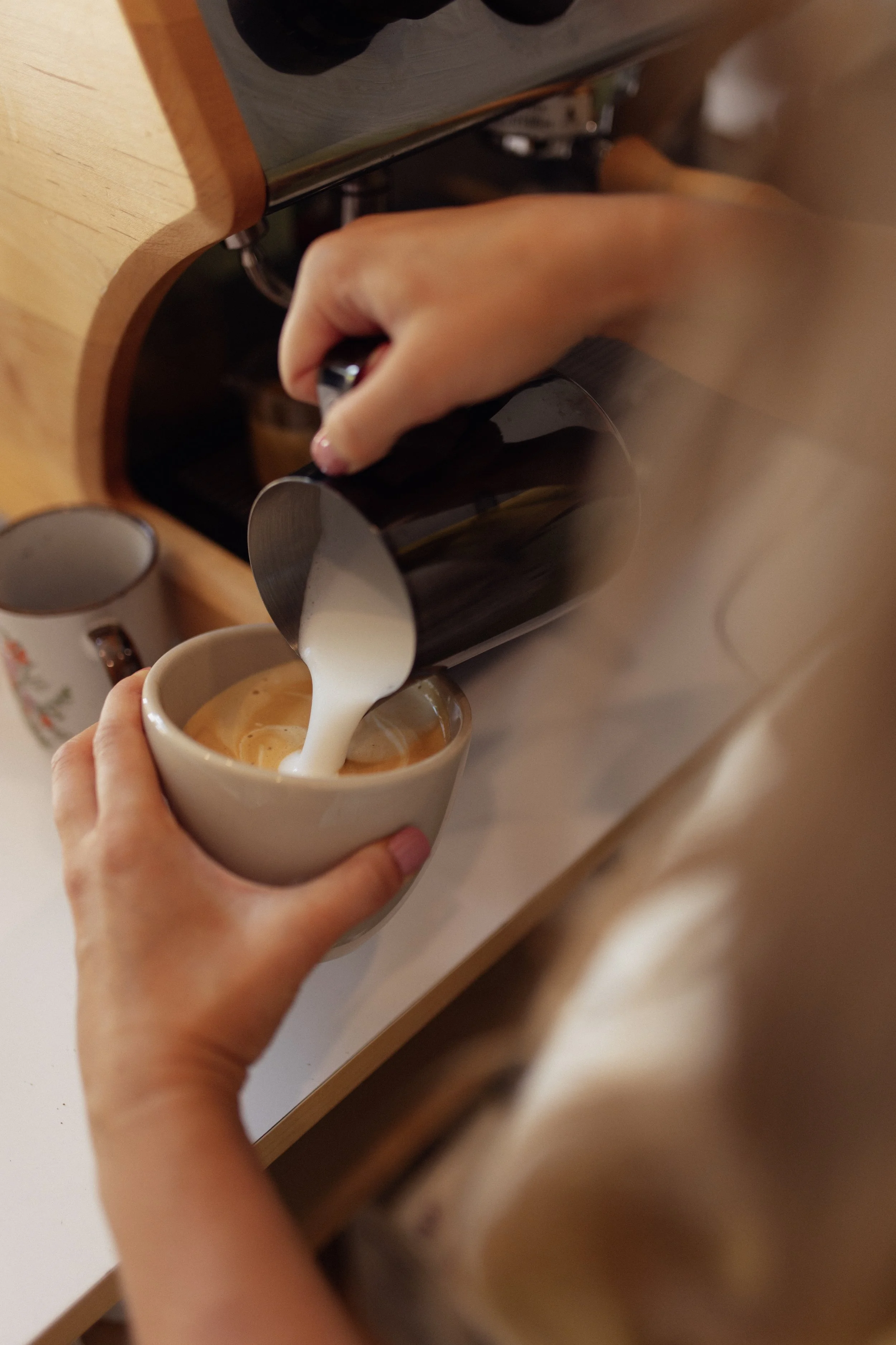 A person pouring steamed milk into a cup of espresso for making a cappuccino or latte. The cup is being held in one hand, and the other hand is holding a milk frothing pitcher. The scene is set on a kitchen countertop with mugs in the background.