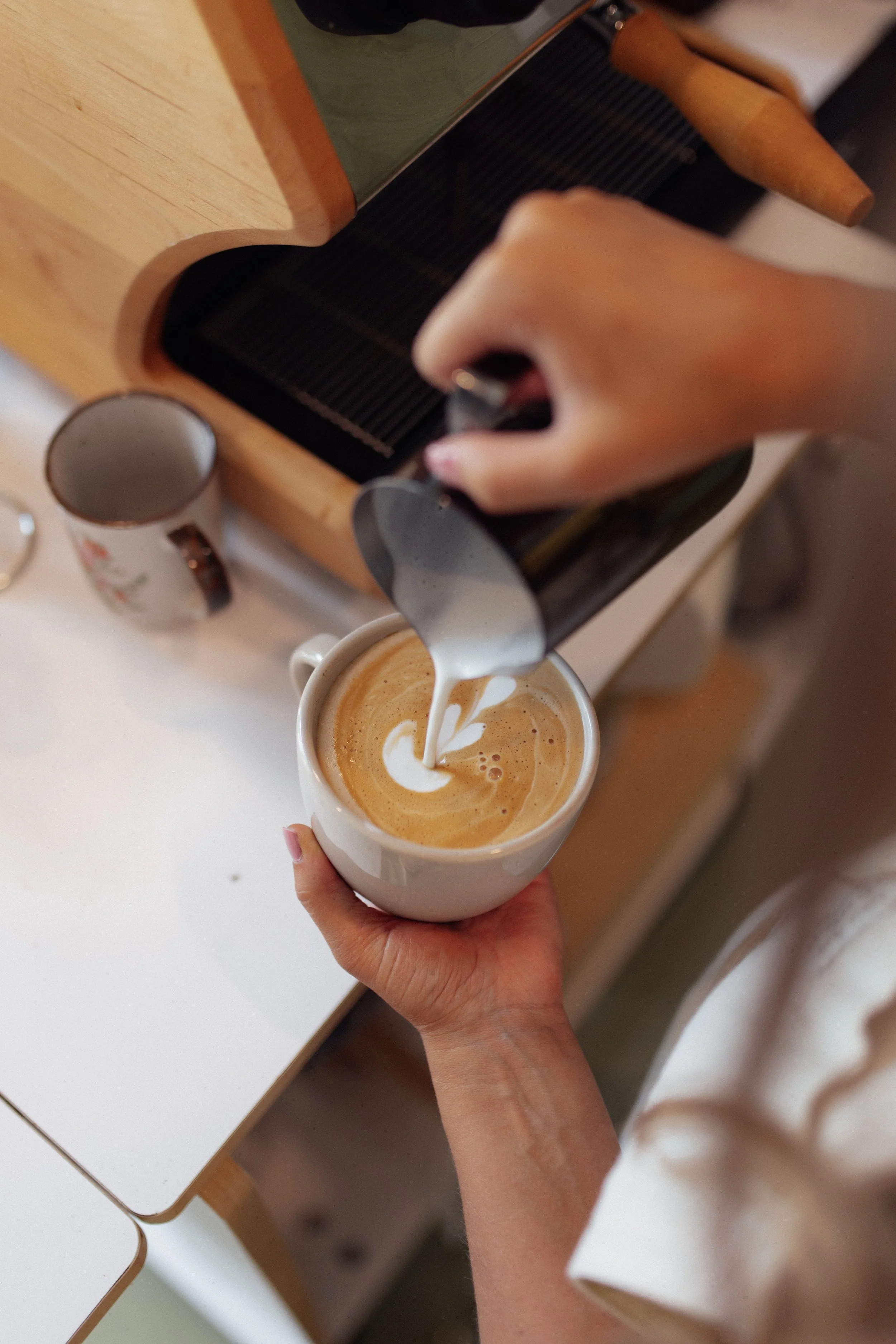 A person is pouring steamed milk into a cup of coffee to create latte art, with a latte mug being held in one hand.