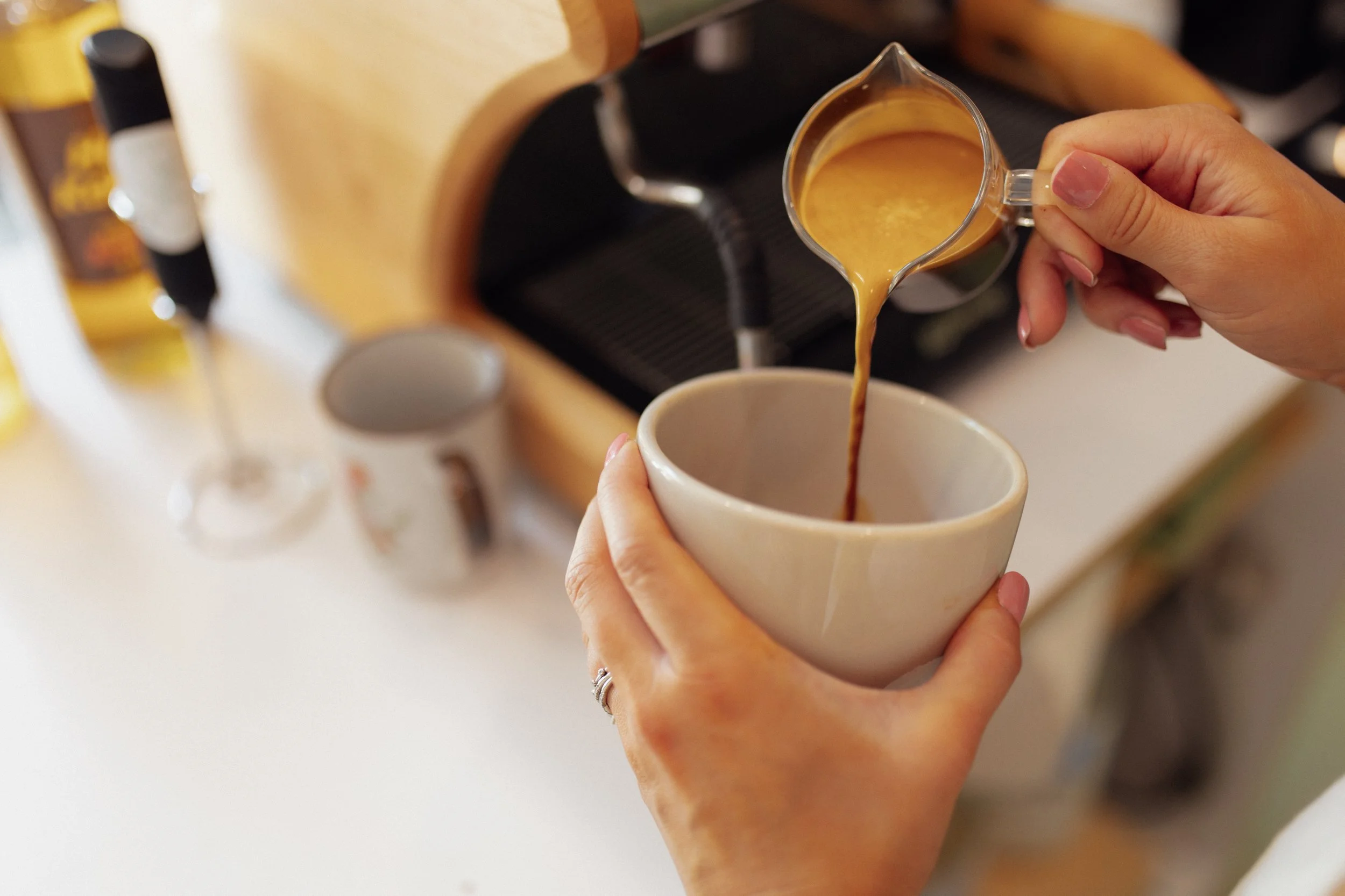 A person pours coffee from a small pitcher into a mug at a coffee shop or kitchen.