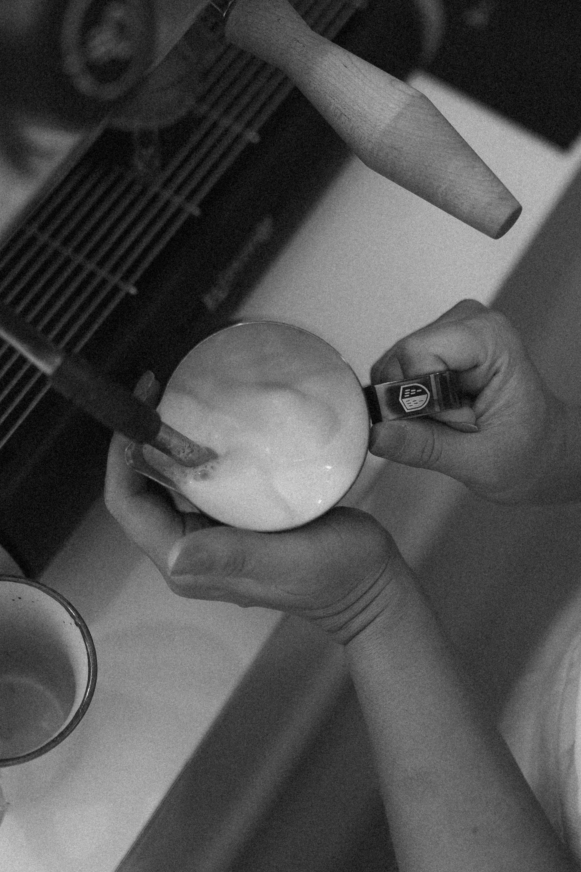 A person holding a white mug with a logo on the handle as they pour a liquid into it. There are cups and a grid-like shelf or rack nearby.