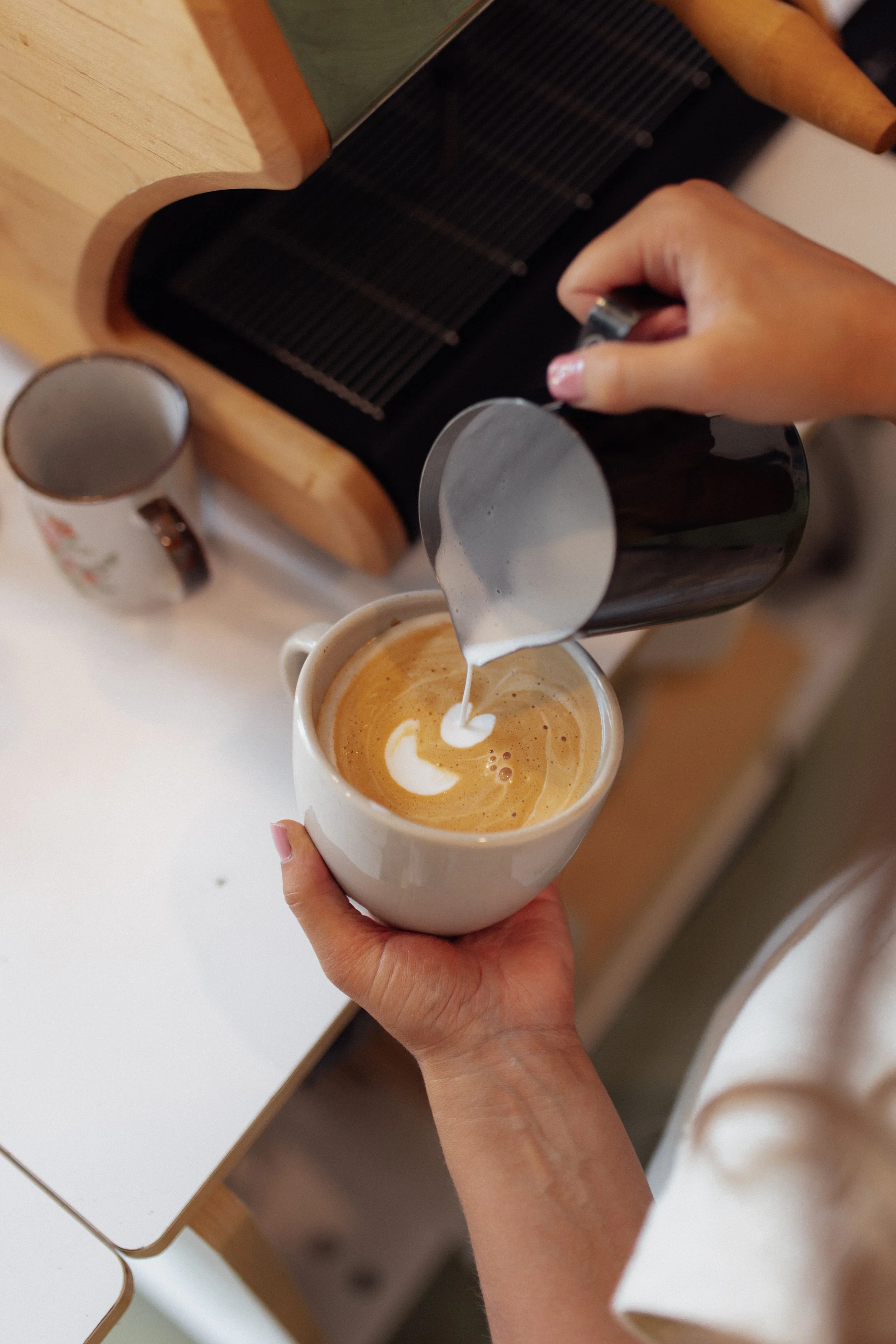 A person holding a white coffee mug with latte art, pouring steamed milk into it, near a black coffee press and a mug on a wooden table.