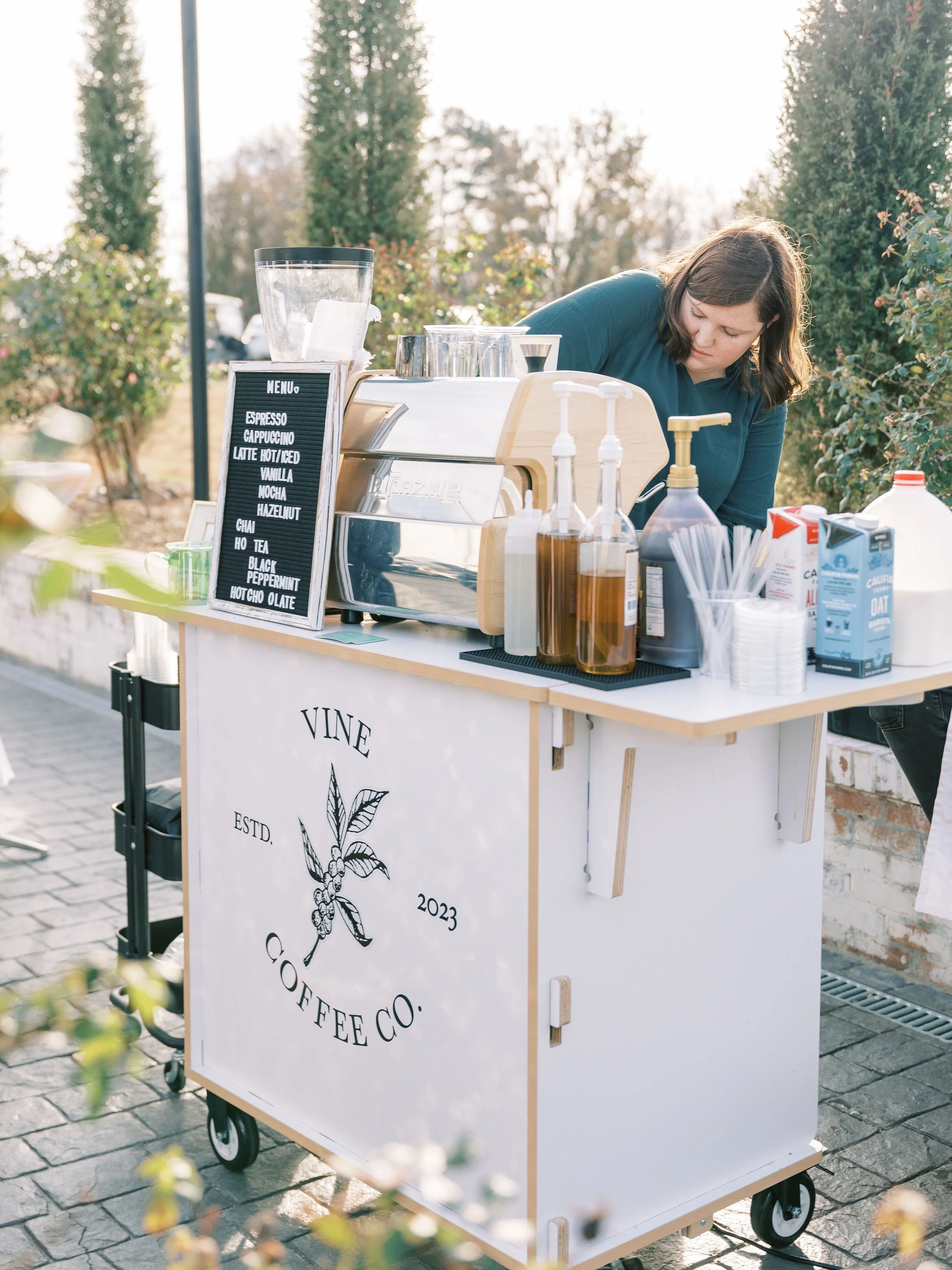 A mobile coffee stand outdoors with a woman preparing drinks. The stand displays a menu with coffee options like espresso and latte, and various bottles and supplies are arranged on the counter. The setup is surrounded by trees and natural daylight.