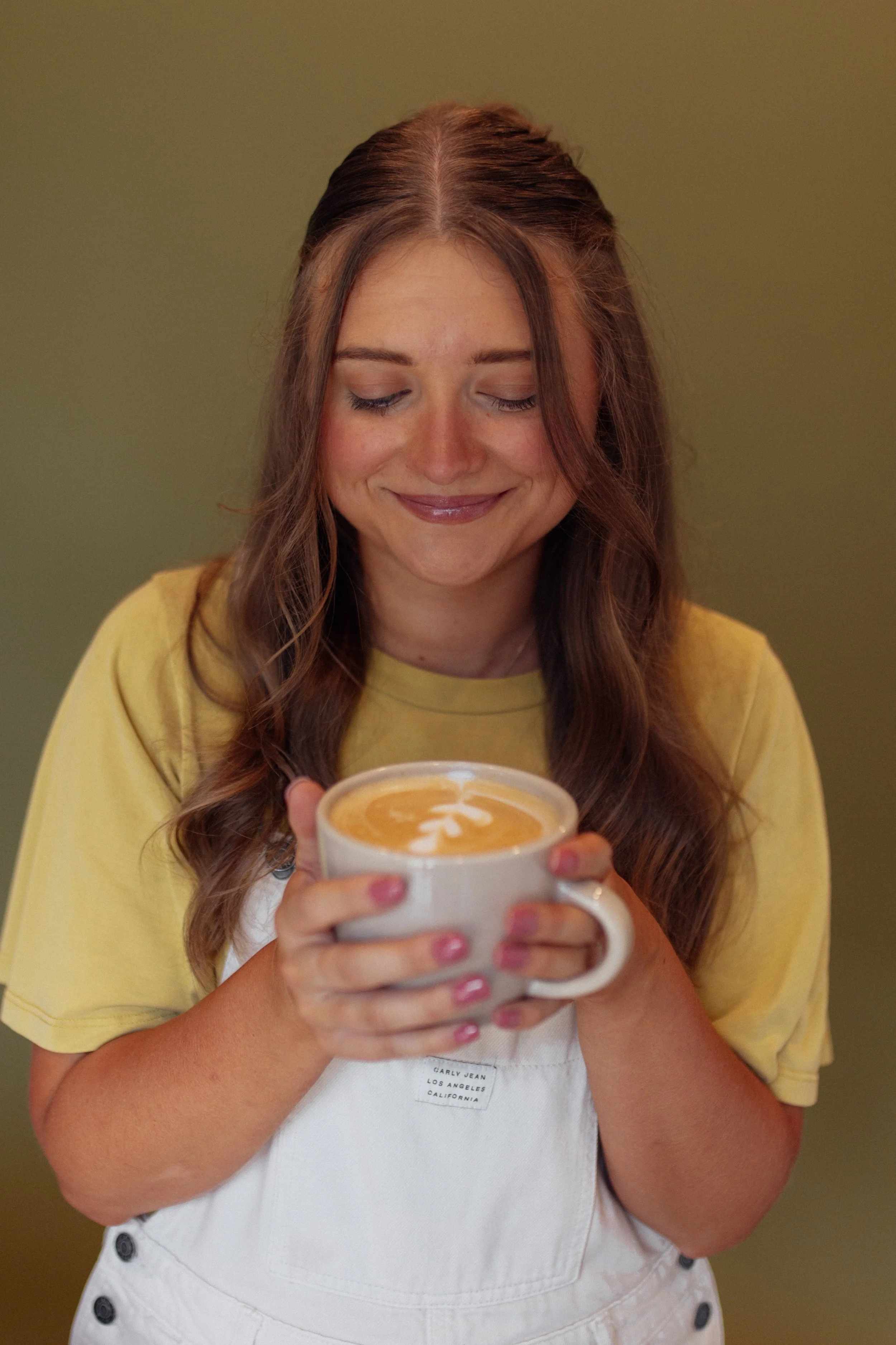 A woman with long brown hair, wearing a yellow T-shirt and a white apron, holding a white mug with a latte art heart design. She is smiling with her eyes closed.