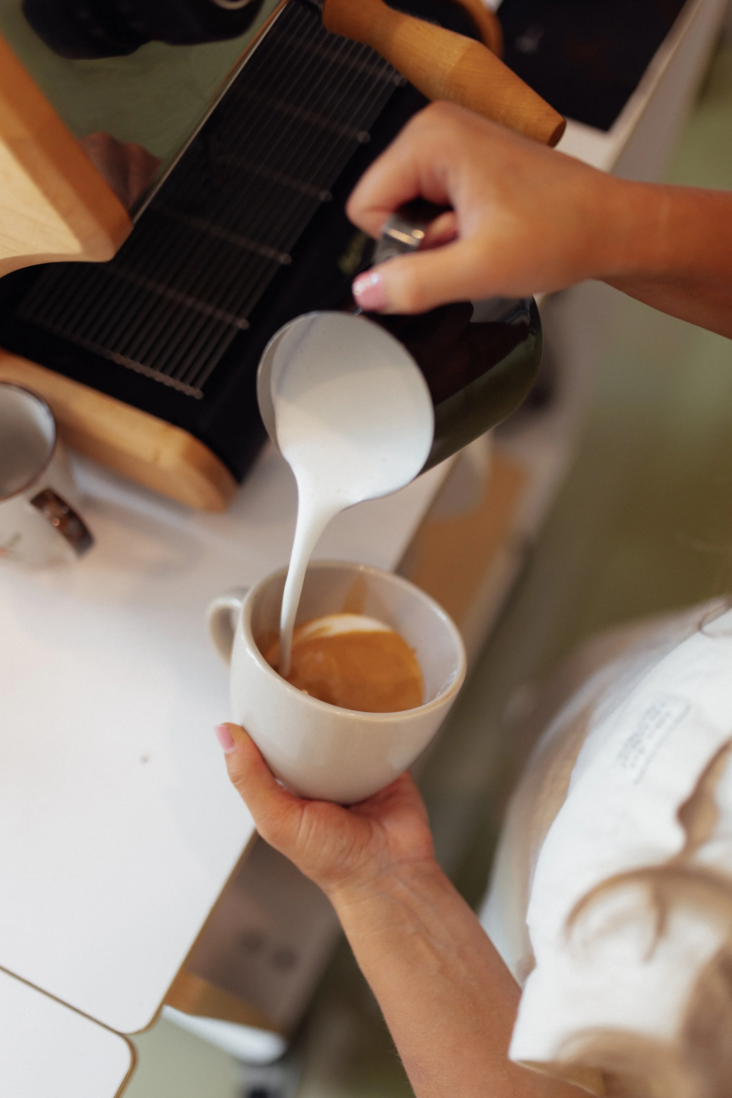 Person pouring milk into a cup of coffee or latte on a white table.