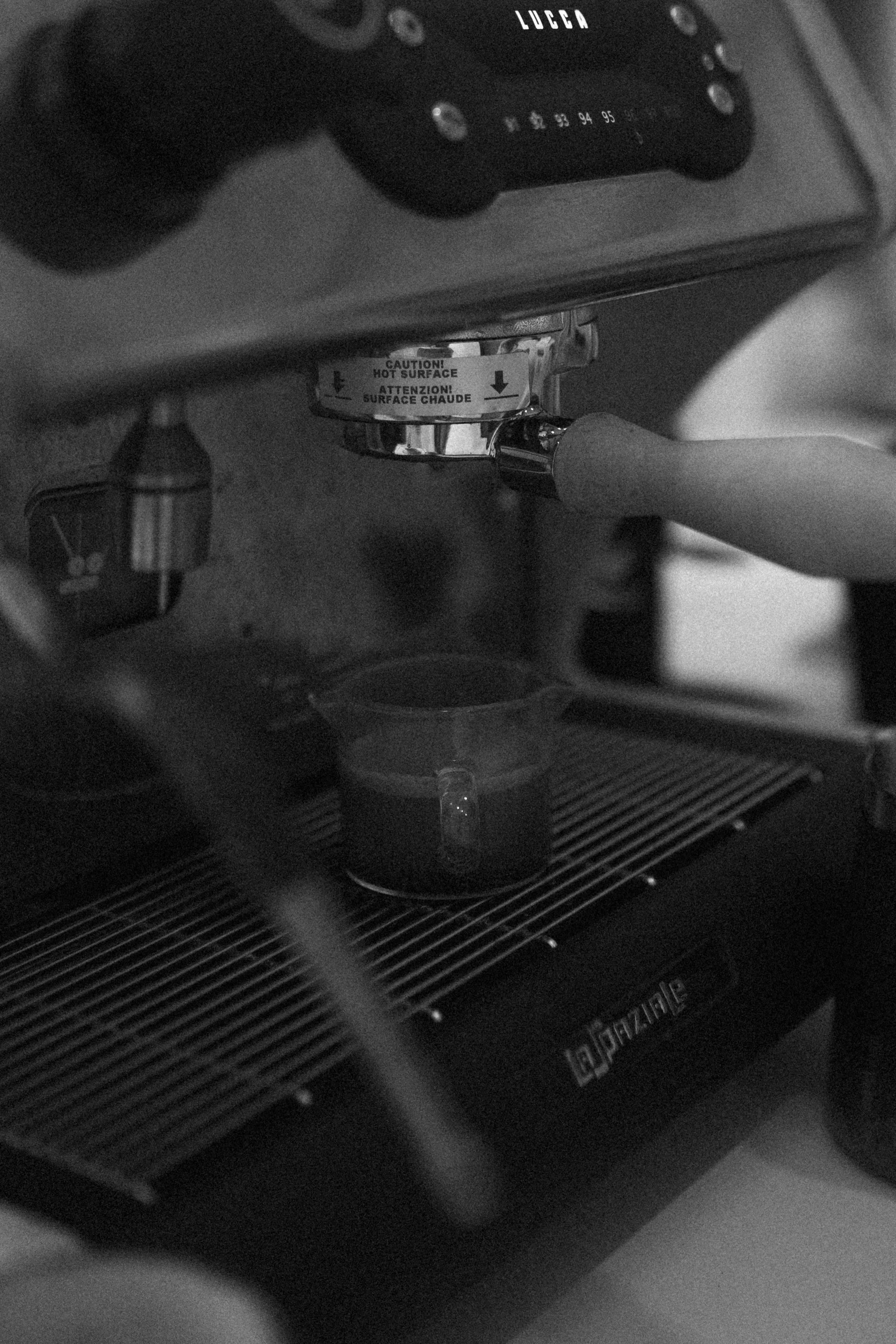Close-up of a coffee machine with a shiny metallic portafilter attached and a caution sticker warning about hot surface. The machine features a control panel with silver knobs, and the text 'LUCCA' is visible on the top.