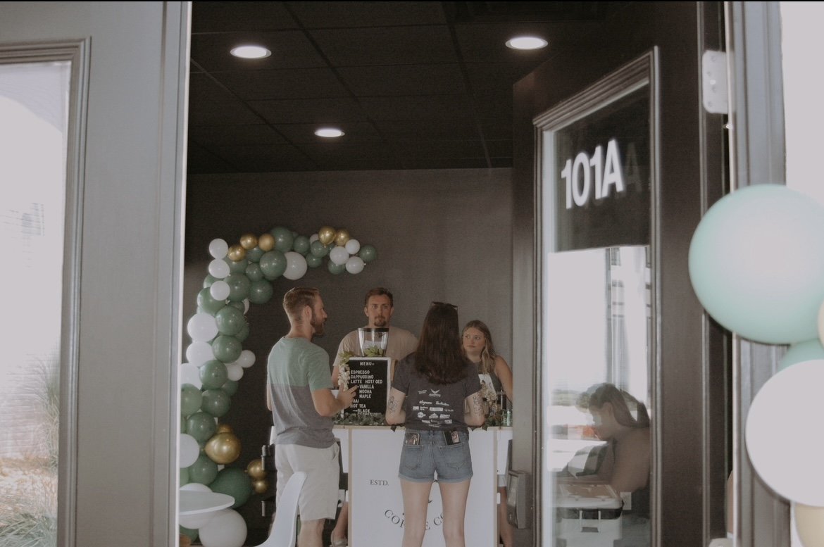 View of a coffee shop interior seen through a glass door with the room number 101A. Inside, a woman with dark hair and denim shorts is talking to a man in a grey t-shirt. Behind the counter, a barista is preparing drinks and a woman is waiting. The b