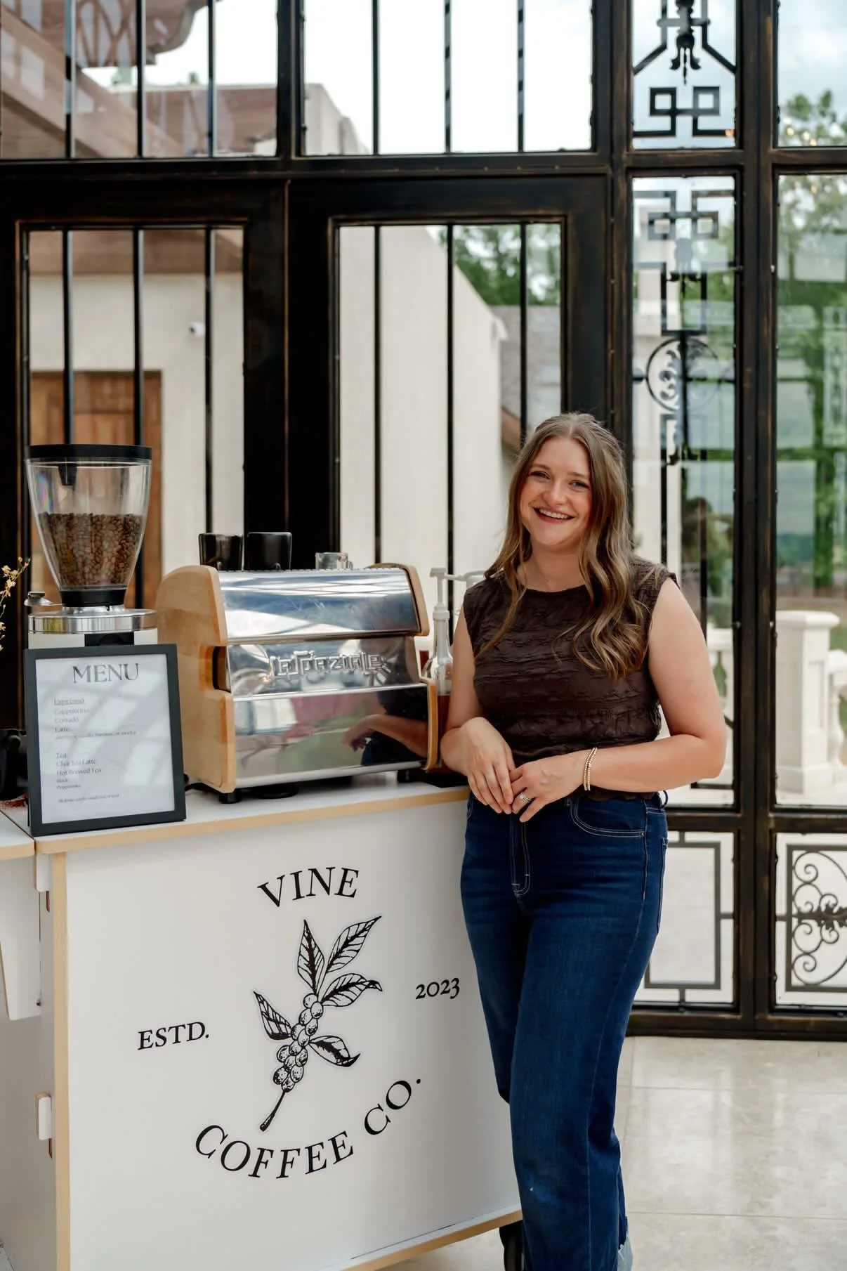 A woman standing next to a coffee stand with a smile, in front of a black metal and glass fence, with coffee equipment and a menu on display.