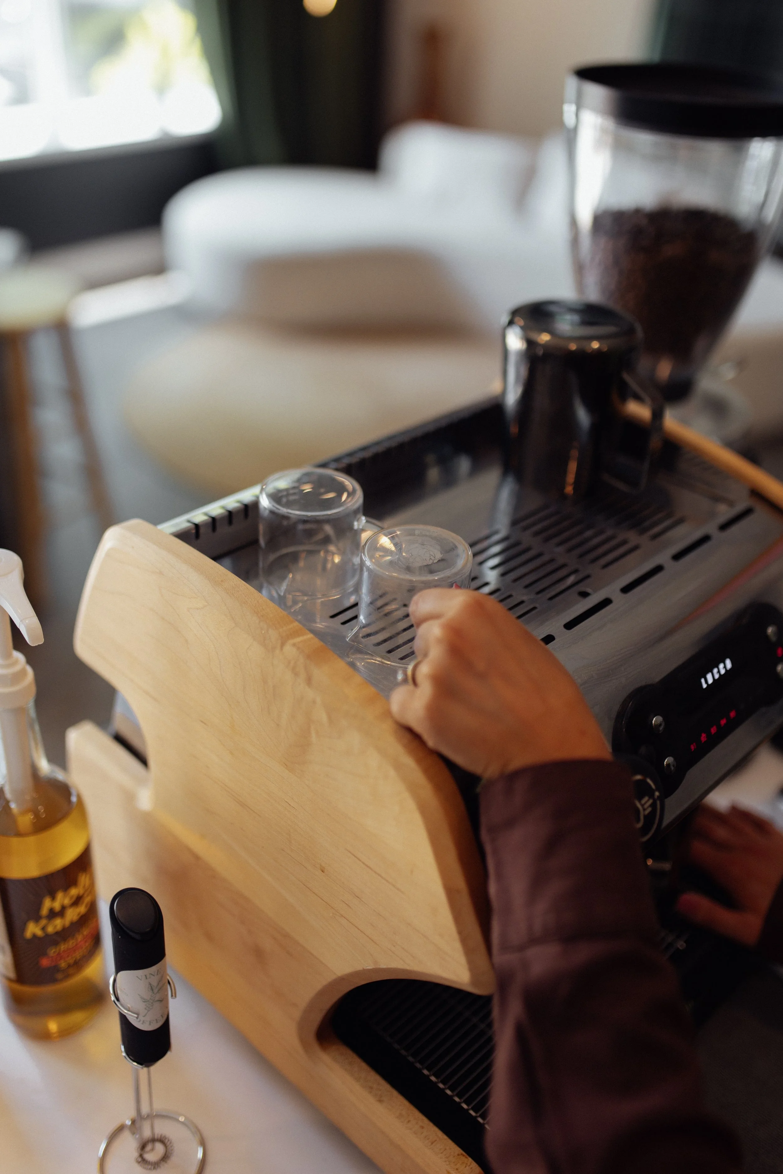 Person pressing down on a coffee machine with a wooden frame, with cups placed underneath, in a kitchen or coffee shop setting.