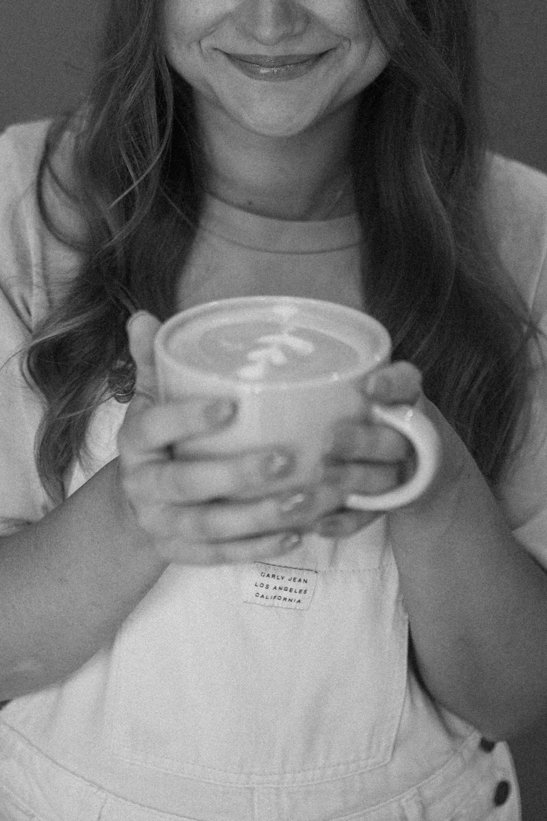 A woman smiling and holding a cup of coffee or tea. She is wearing a light-colored shirt with a visible tag that reads "Clarify Jean Los Angeles California." Her long hair is wavy.