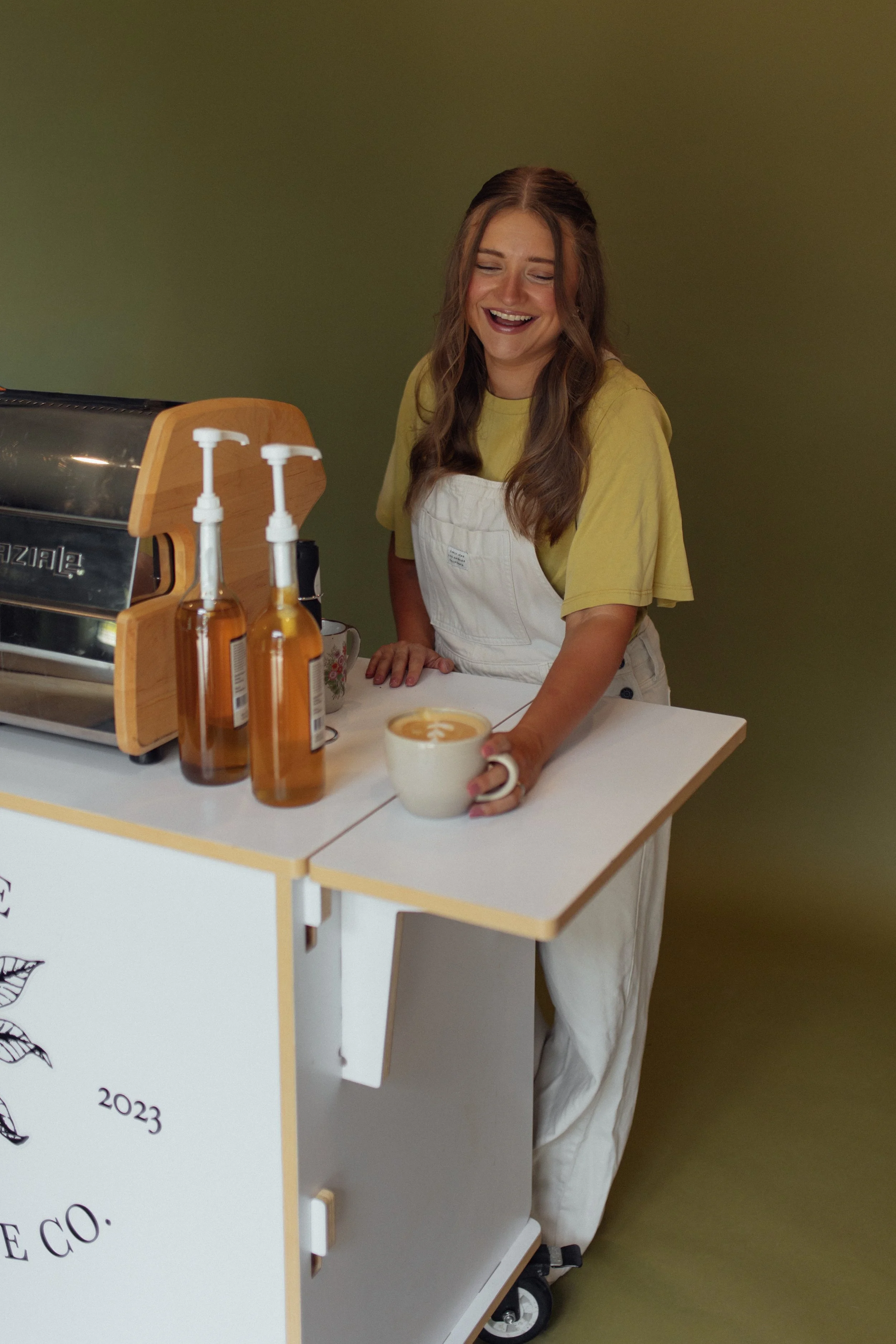 Woman in yellow shirt and white apron smiling, holding a cup of coffee or latte with latte art, standing behind a white counter with bottles of hand soap or sanitizer, an espresso machine, and decorative plants.