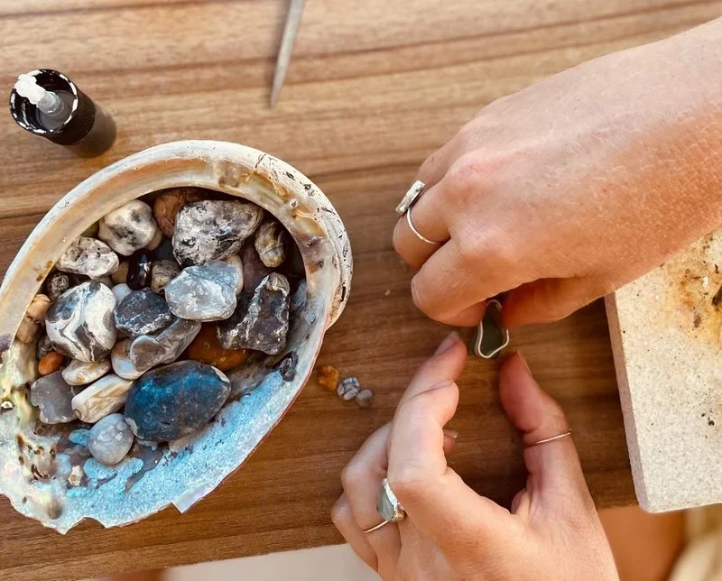 Hands working with jewelry and a collection of stones on a wooden surface.