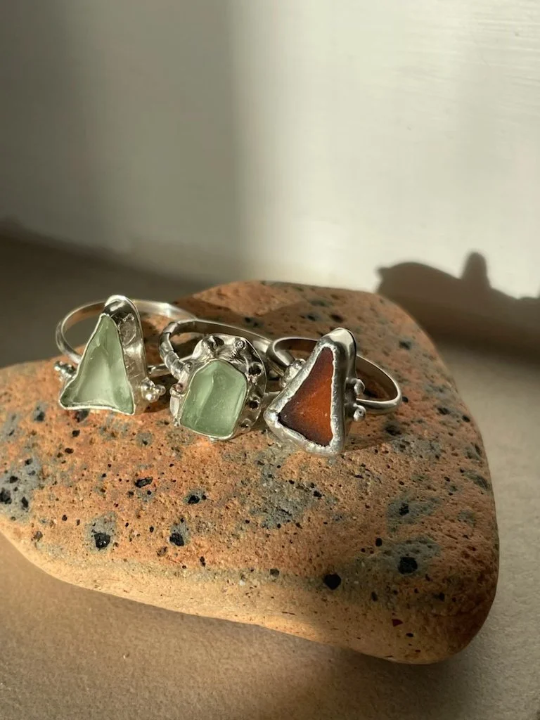 Three silver rings with colored stone insets rest on a textured brown stone surface, with natural light casting shadows.