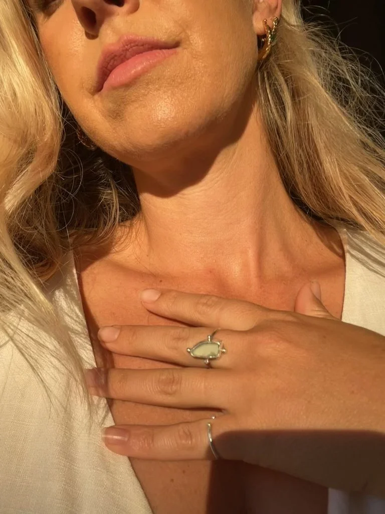 Close-up of a woman's neck and hand, her hand resting on her chest with a large, light-colored stone ring and a simple band ring. She has blonde hair, earrings, and is wearing a light-colored top.