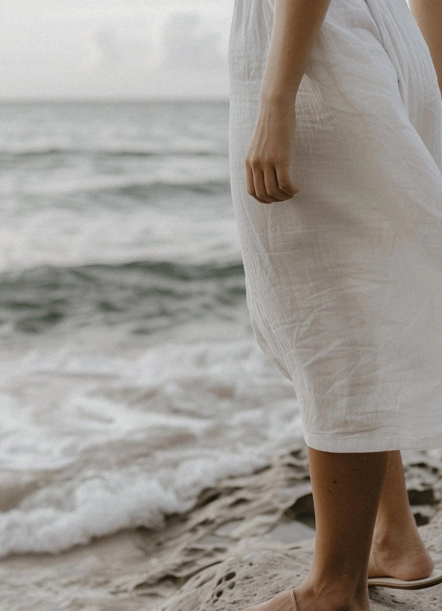 Close-up of a person's hand and lower body standing on a sandy beach near the ocean, wearing white linen clothing.