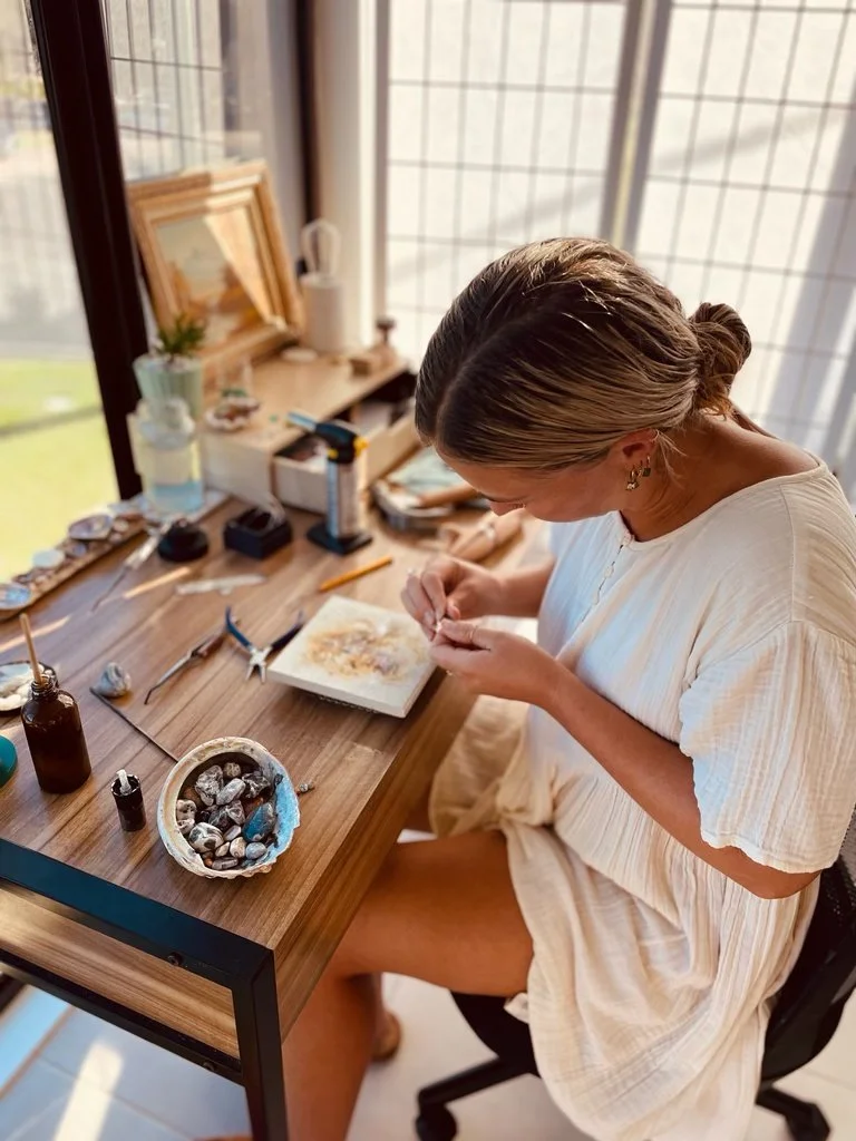 Woman working on jewelry at a wooden desk, with various tools and stones, in front of a bright window.