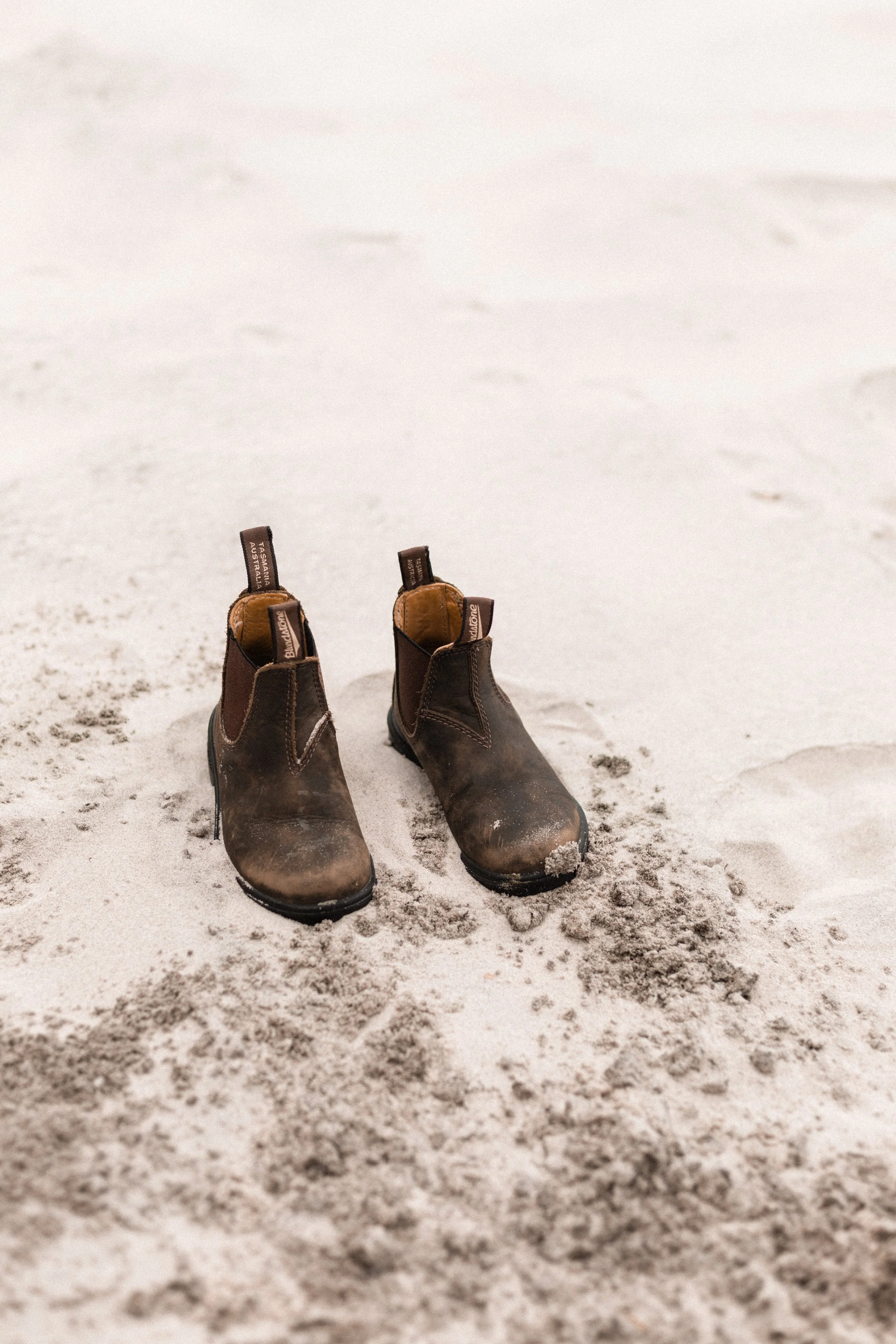 Pair of brown work boots on sandy ground with footprints.