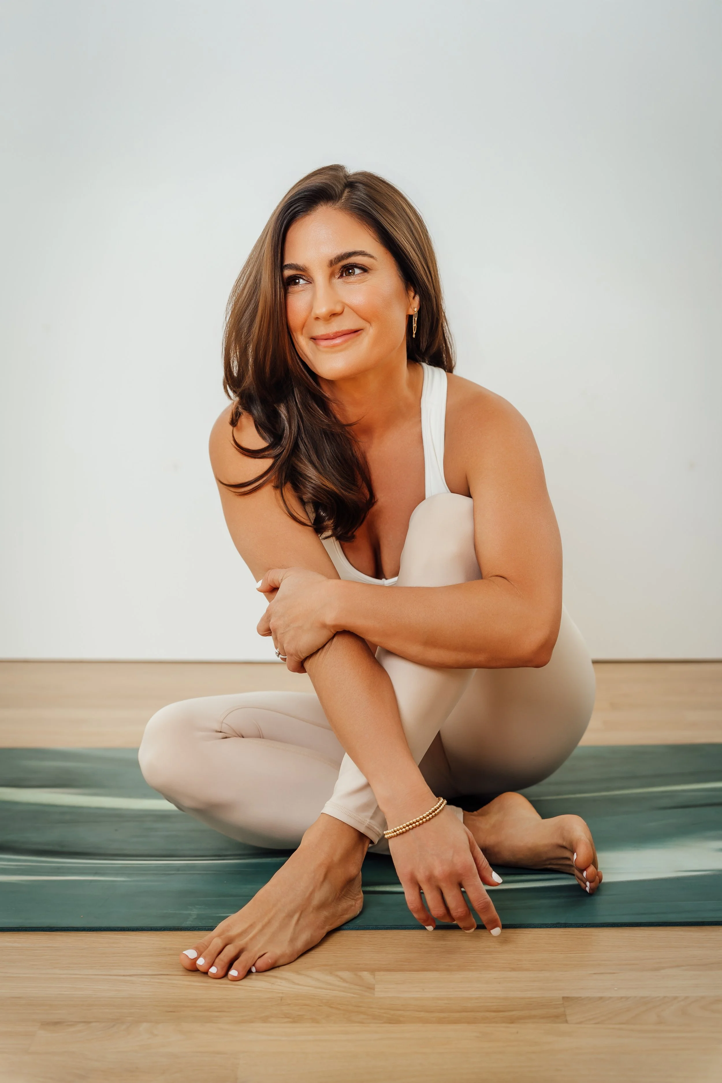 A woman practicing yoga on a mat, sitting cross-legged, with a calm and focused expression, against a plain white wall.