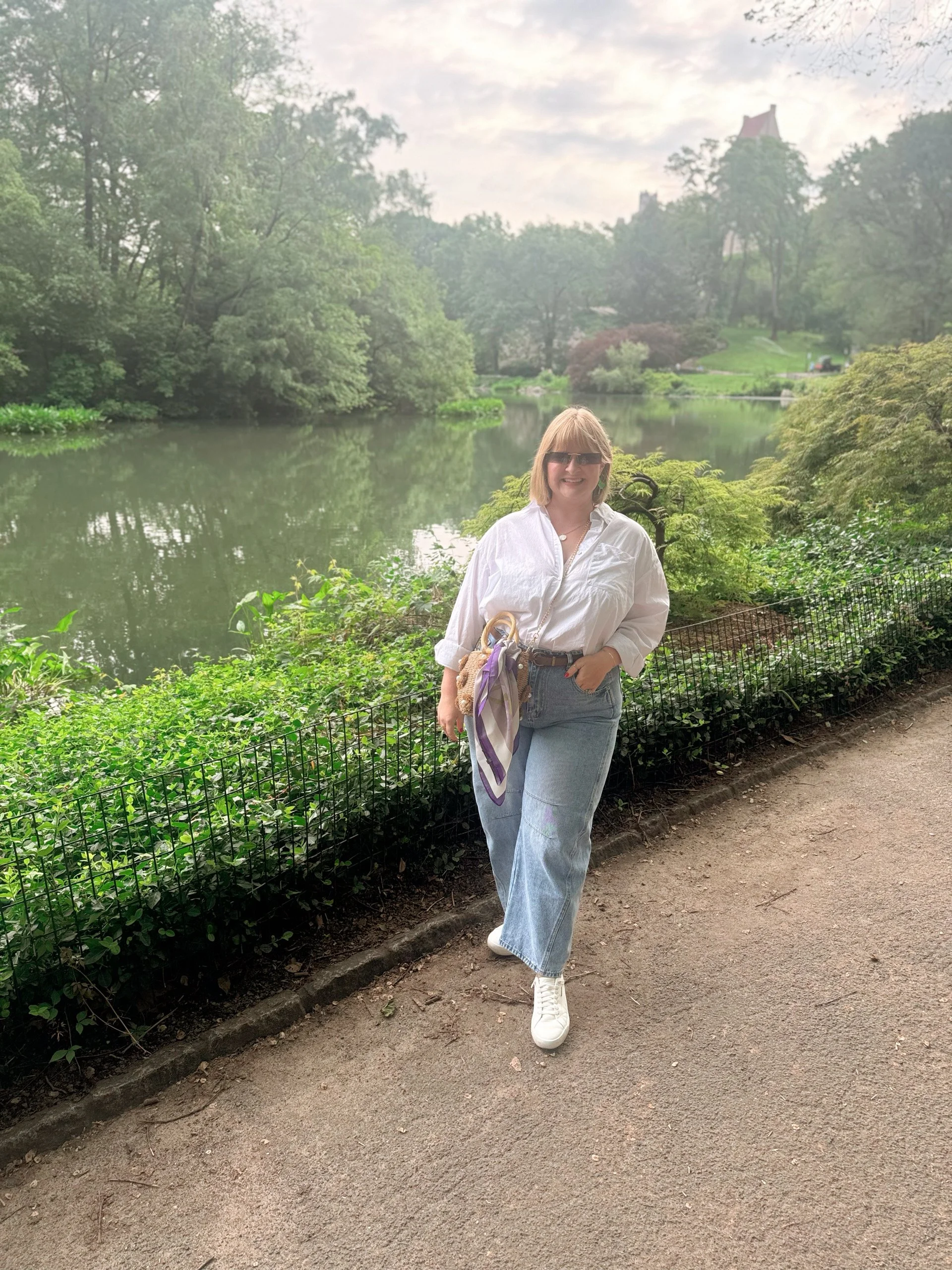A woman with blonde hair wearing sunglasses, a white shirt, and light blue jeans, smiling and standing on a dirt path by a body of water, surrounded by green trees and foliage.