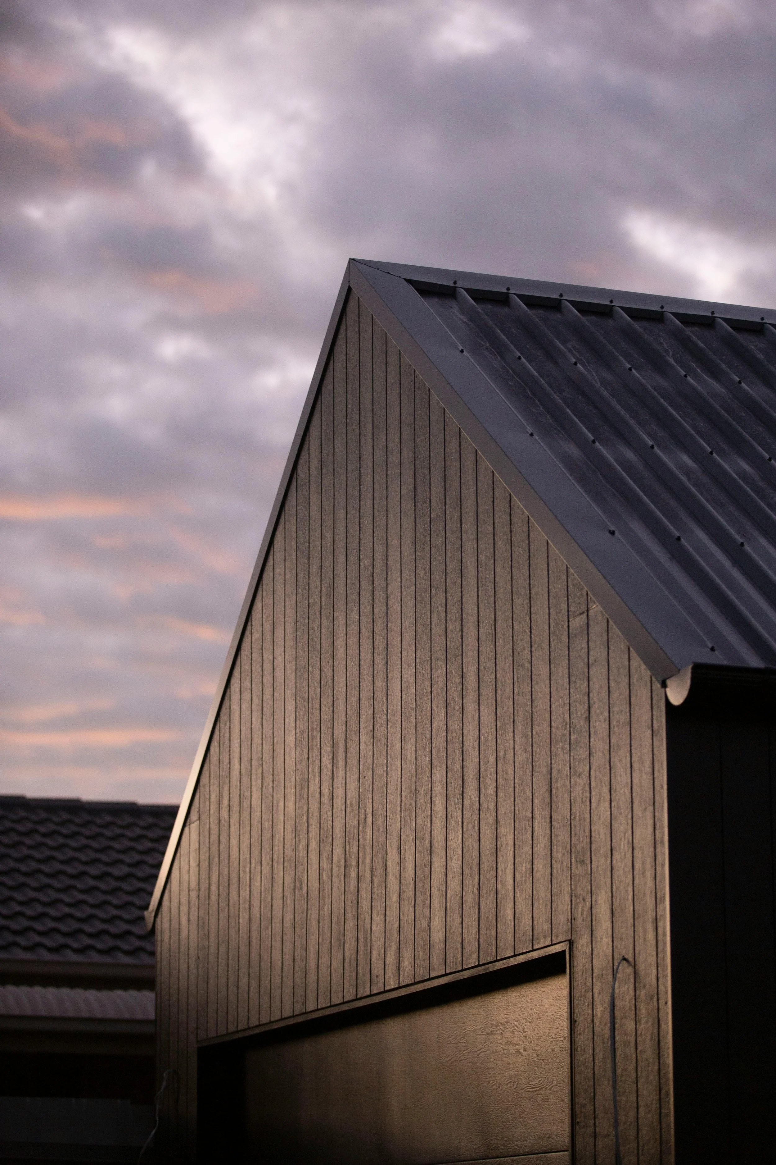 Close-up view of a house with vertical wooden siding and a metal roof, under a cloudy sky at sunset.