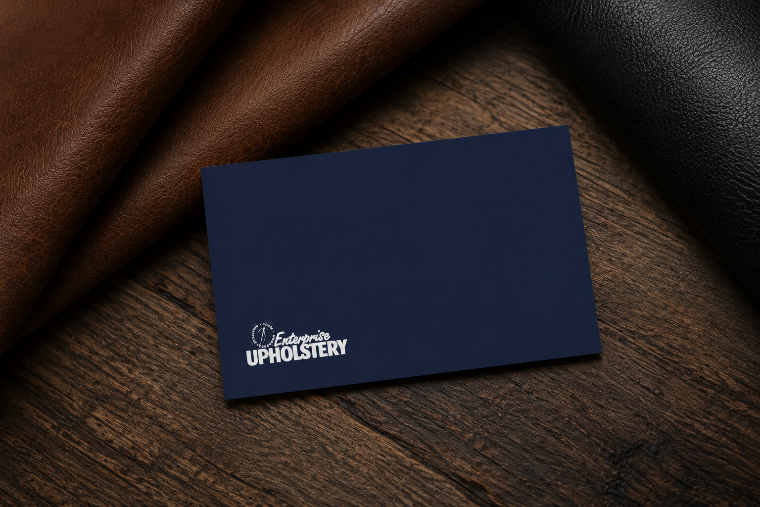 A dark blue business card with 'Enterprise Upholstery' written on it in white, placed on a wooden surface, with rolls of brown and black leather around it.