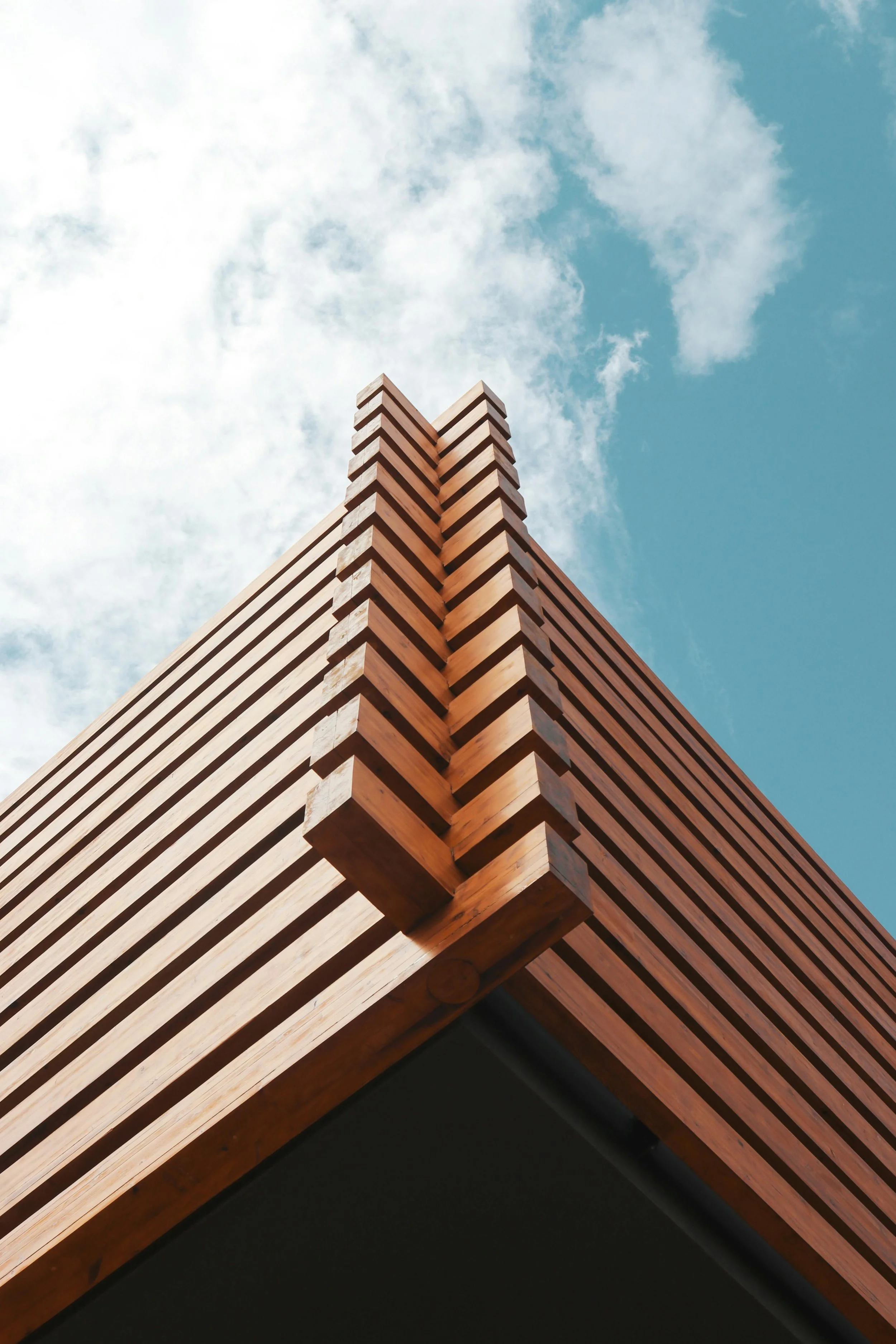 Close-up of a corner of a modern building with wooden slats and a black facade at the bottom, under a partly cloudy sky.
