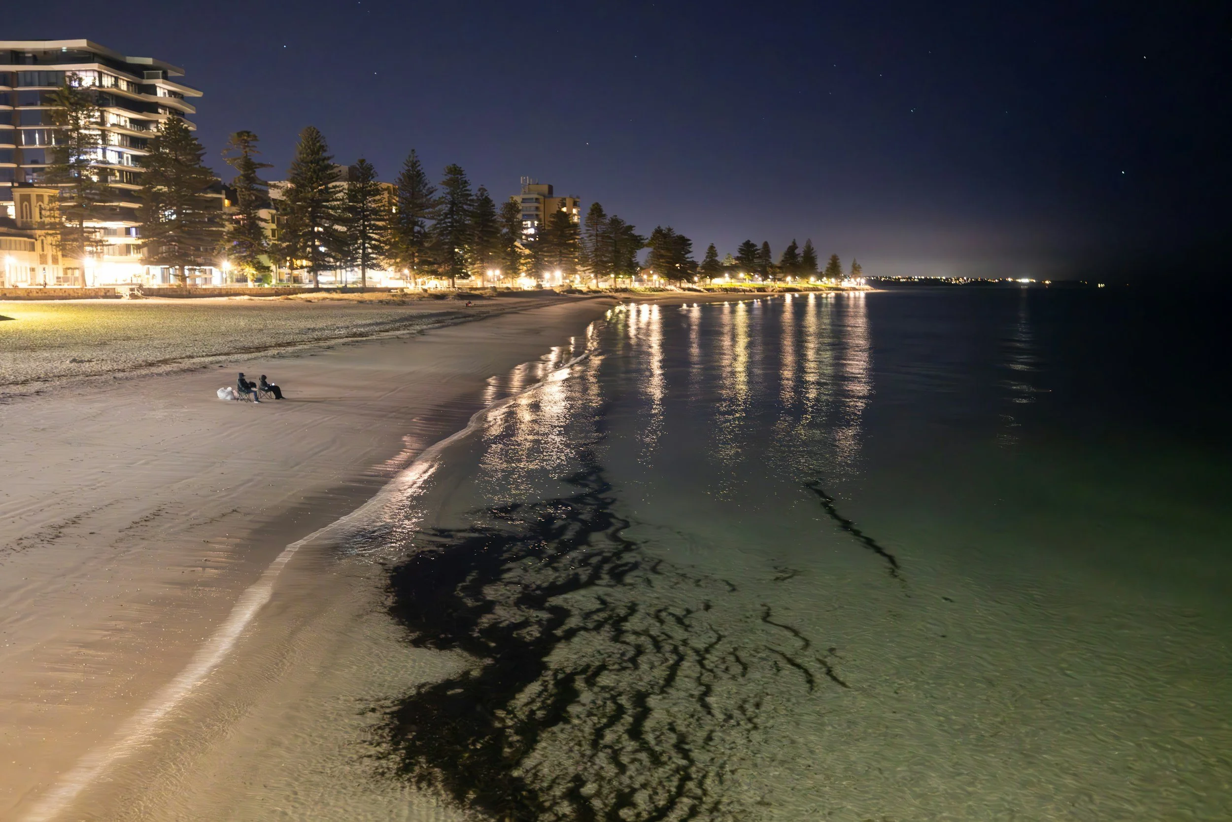 Nighttime view of a beach with calm water, two people sitting on chairs near the shore, multi-story beachfront buildings with lights, and a clear dark sky with stars.