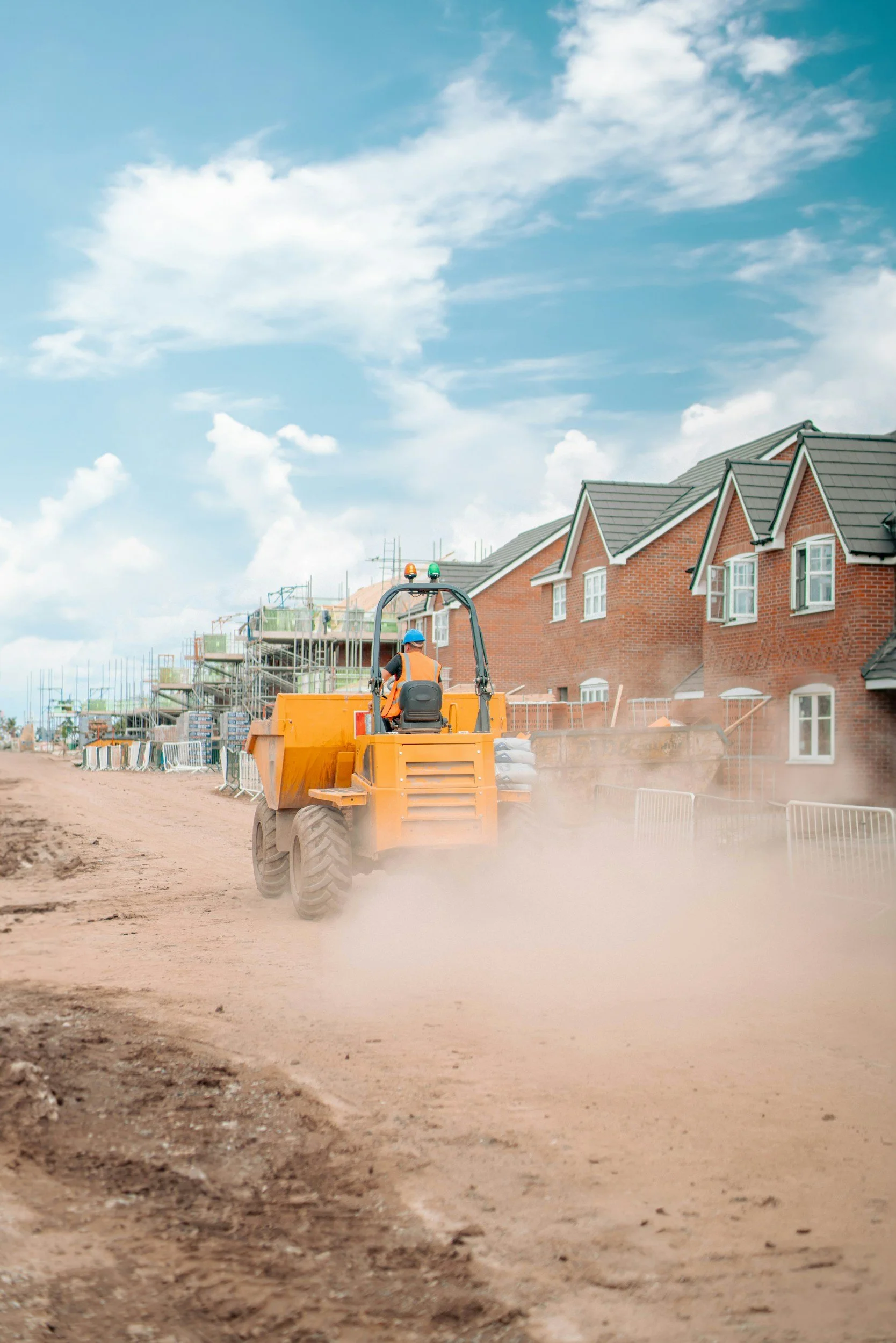 Construction worker operating a yellow road roller on a dirt road with houses under construction and blue sky with clouds in the background.