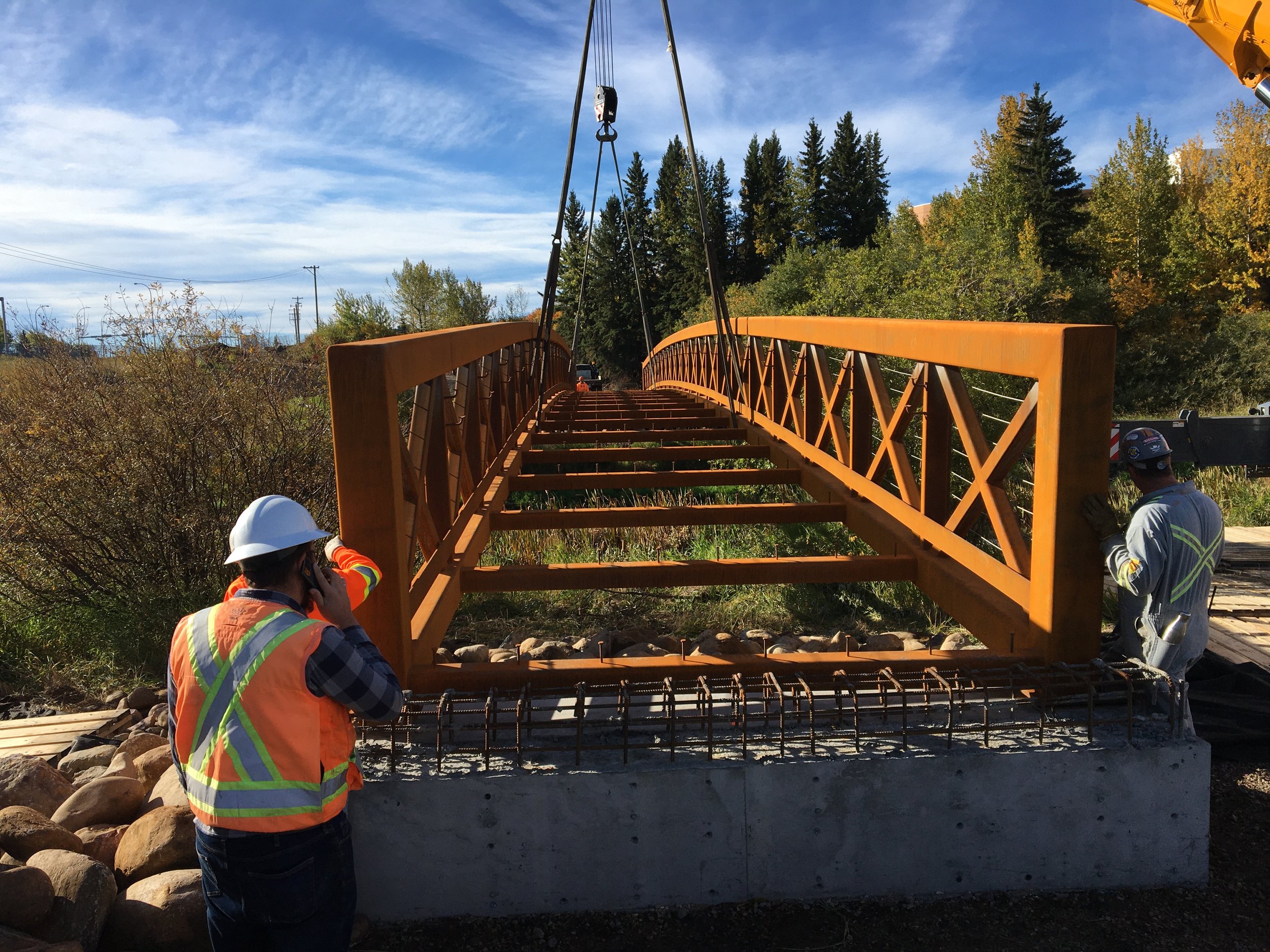 Construction workers in safety gear installing a steel bridge over a rocky area with trees and a blue sky in the background.