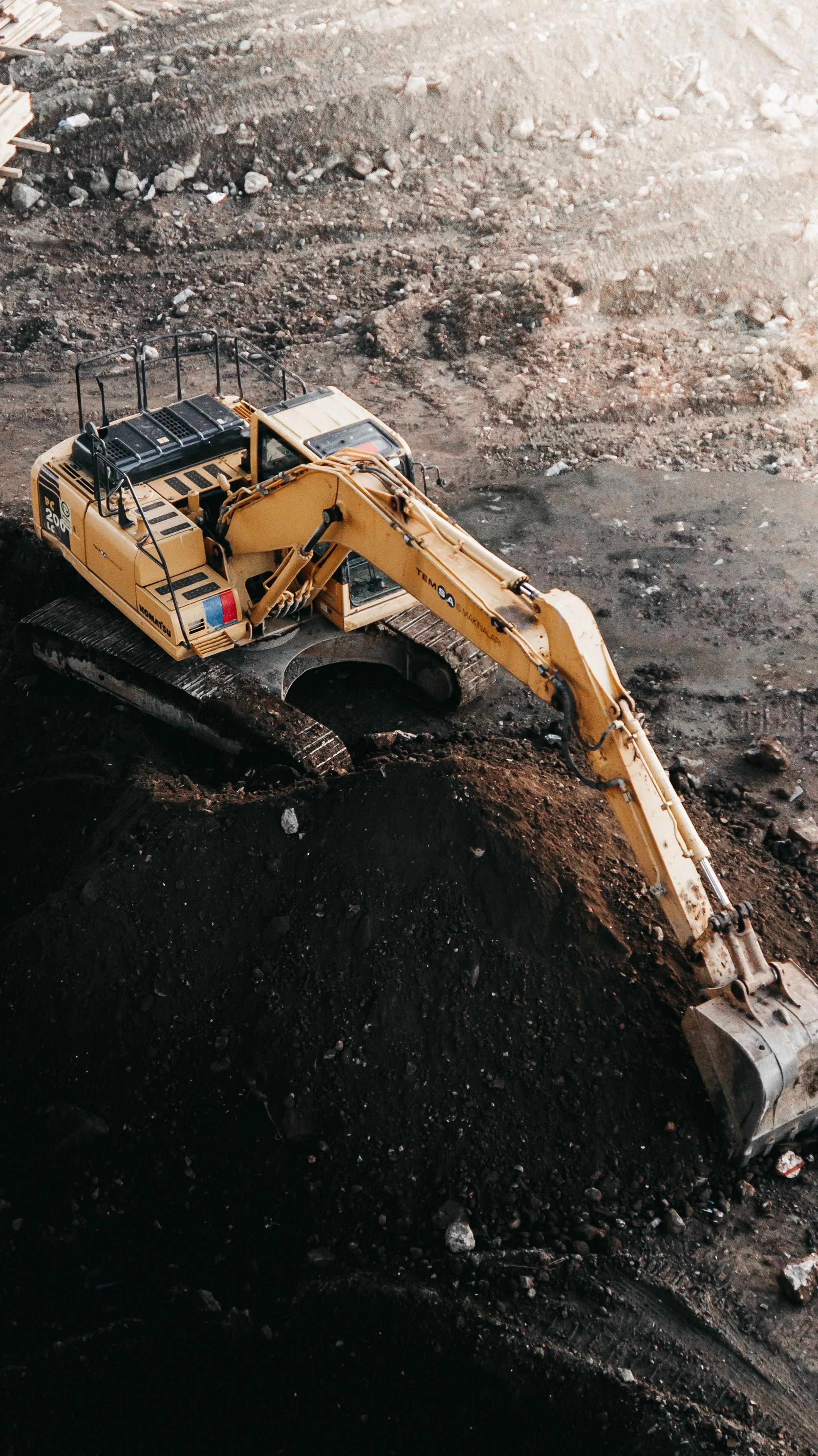 A yellow excavator working on a construction site, digging into the dark soil with rocks scattered around.