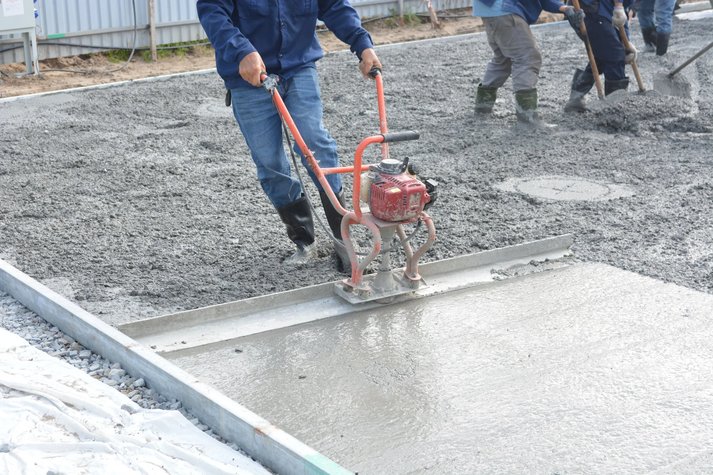 Workers are pouring and leveling concrete on a construction site, wearing gloves and rubber boots.