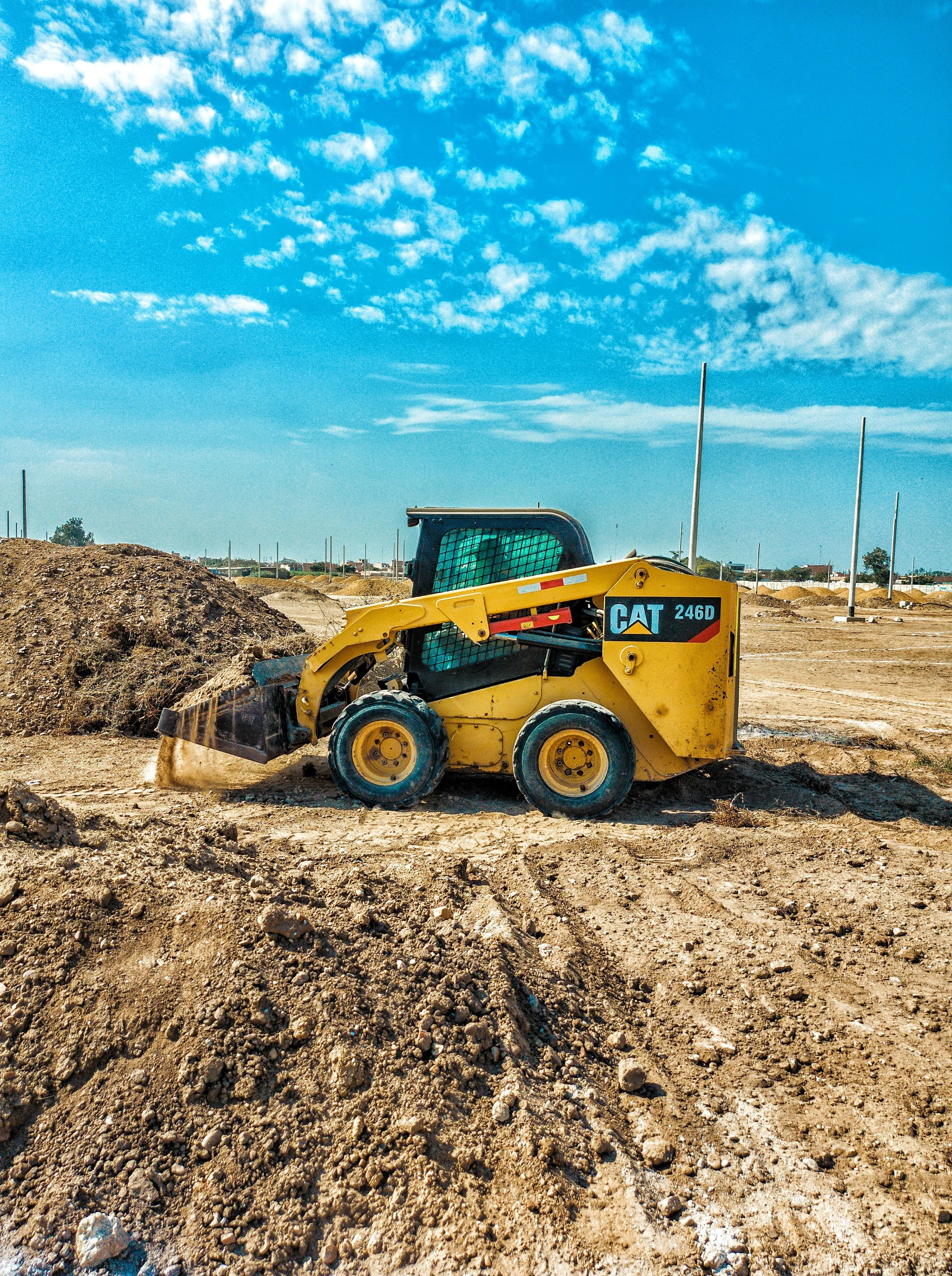 Construction site with a small yellow Caterpillar skid steer loader moving dirt and rocks under a blue sky with scattered clouds.