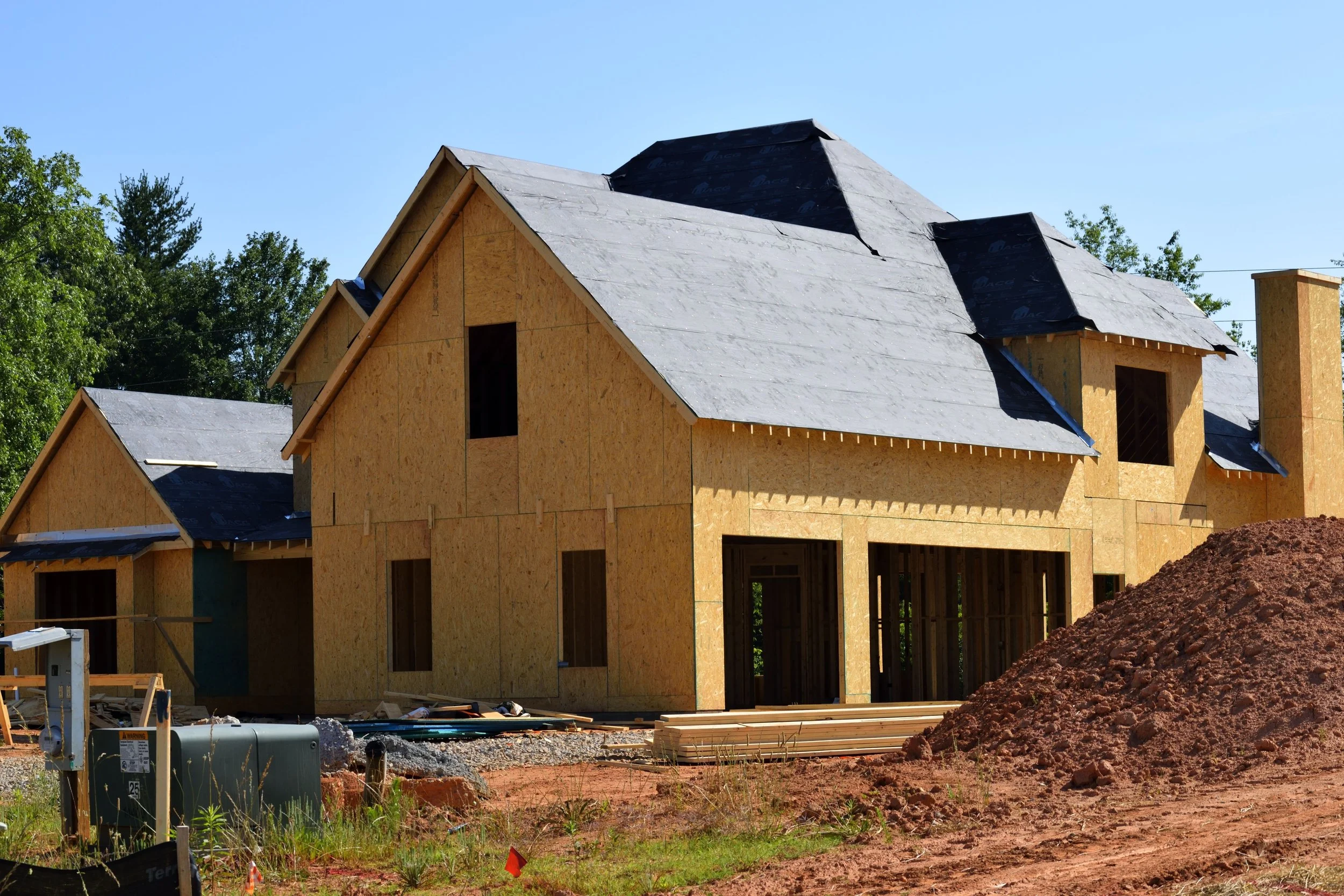 A house under construction with a wooden frame and roof in progress, surrounded by building materials and dirt.