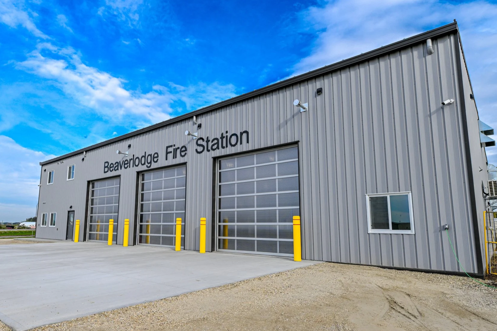 Metal building labeled "Beaverlodge Fire Station" with large garage doors, small windows, and yellow safety bollards in front, under a partly cloudy blue sky.