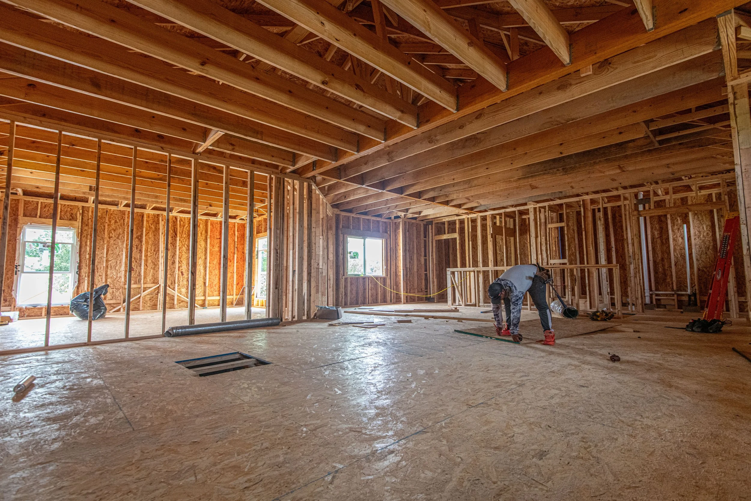 Interior view of a house under construction with exposed wooden framing, large windows, and two workers measuring the floor.
