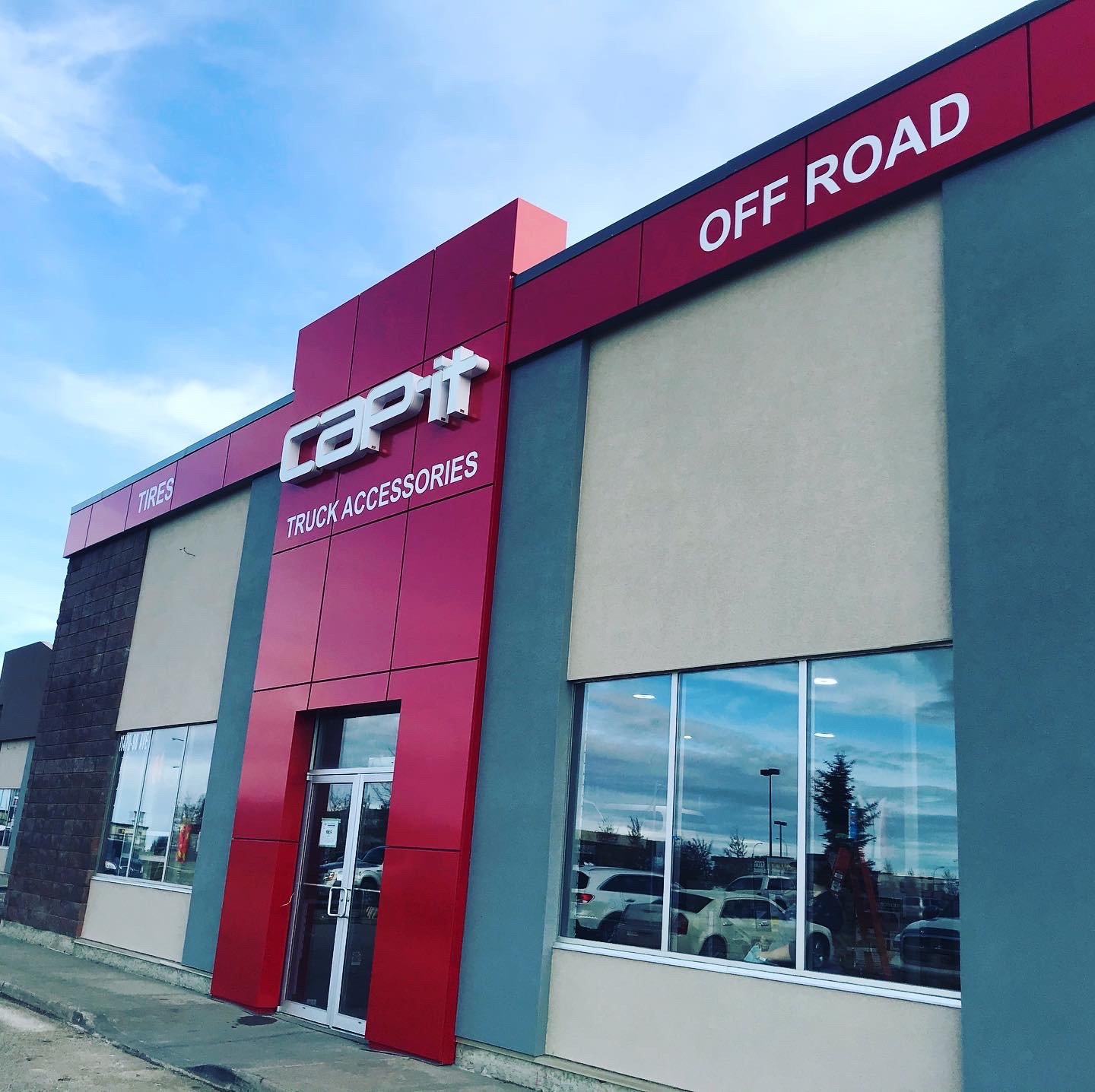 Exterior of a store with red and beige walls, large glass windows, and signage for 'Capat Truck Accessories,' advertising tires and off-road products, located in a shopping area with parking and trees reflected in the windows.