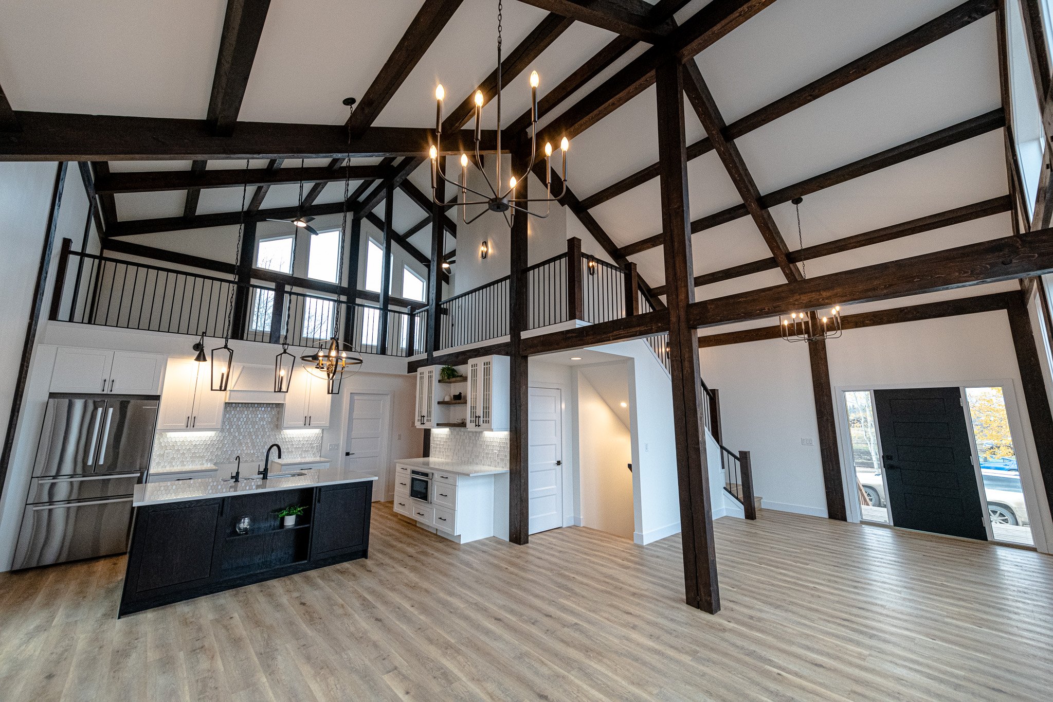 Open-concept kitchen with white cabinets, a black island, stainless steel appliances, wooden floors, and a loft area with dark wooden beams and ceiling fixtures.